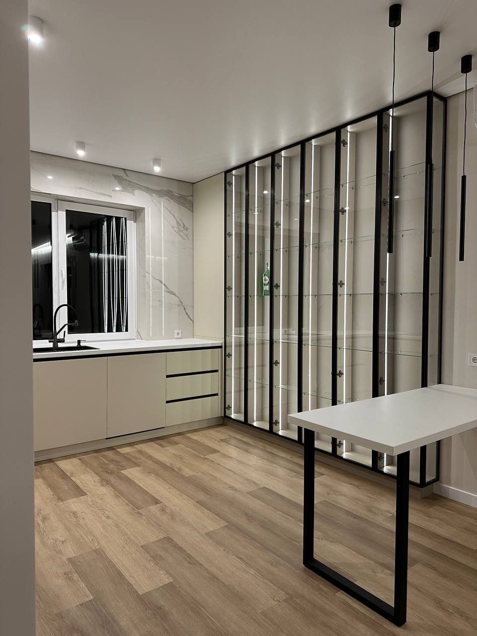 Nowoczesna kuchnia z drewnianą podłogą, jasnymi szafkami i wysoką, przeszkloną witryną z czarnymi ramami i podświetleniem LED. Minimalistyczny stół z czarnymi nogami.