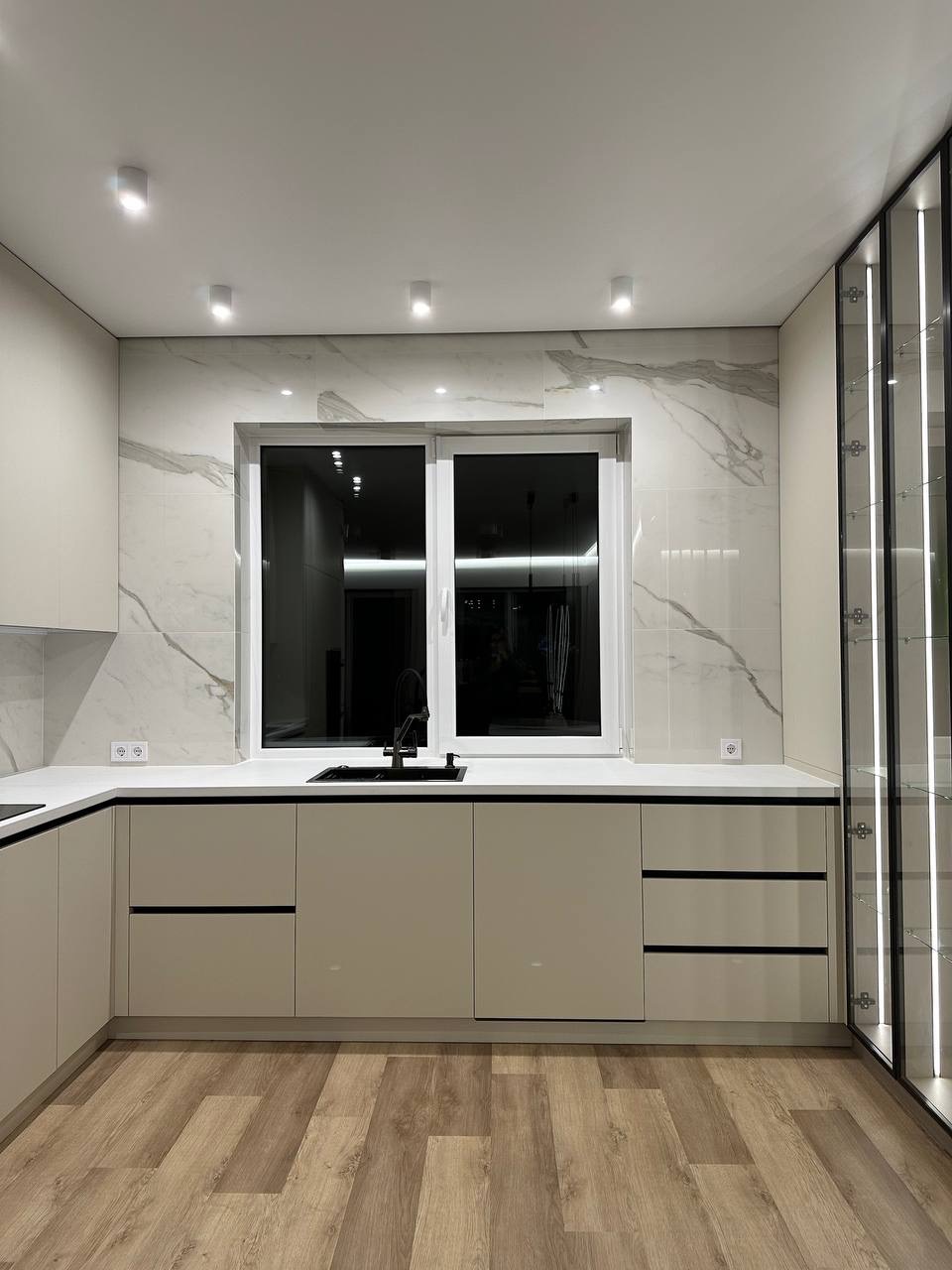 Nowoczesna kuchnia z beżowymi szafkami, marmurowym blatem i ścianą. Czarne akcenty: bateria, listwy, okno. Podłoga drewnopodobna. Szklana witryna z oświetleniem.