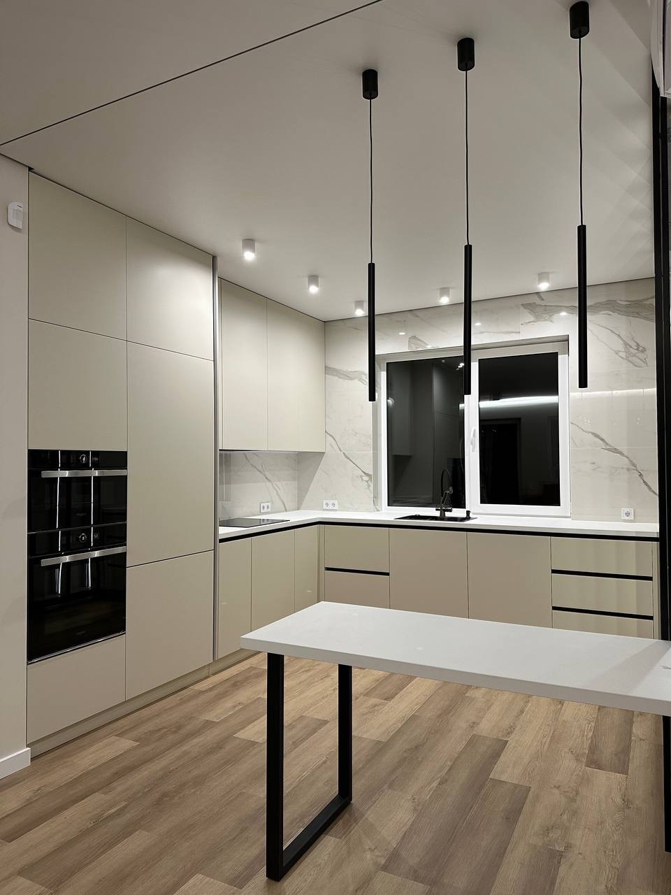 Nowoczesna kuchnia w beżach z marmurowym akcentem, oświetlona wiszącymi lampami. Minimalistyczny stół z czarnymi nogami. Podłoga imitująca drewno.