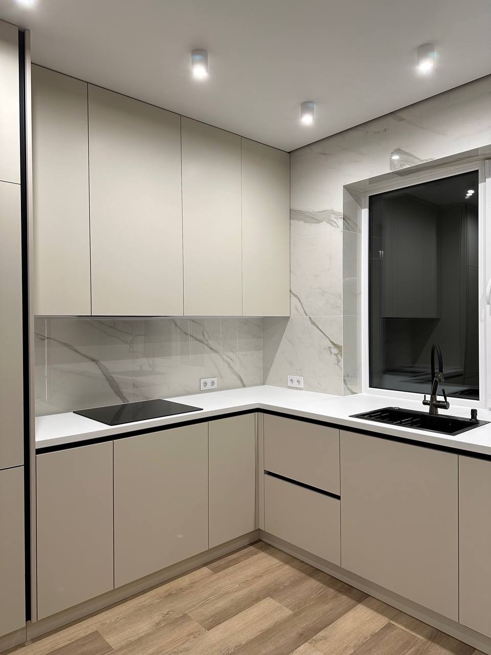 Nowoczesna kuchnia w beżowych tonacjach z marmurowym backsplash'em i czarną armaturą, oświetlona punktowymi lampami sufitowymi, z drewnianą podłogą.