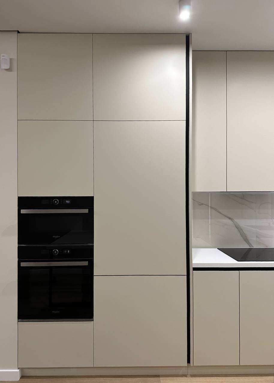 Minimalistyczna zabudowa kuchenna w odcieniach beżu z czarnymi piekarnikami i płytą grzewczą. Gładkie fronty szafek, marmurowe wykończenie nad blatem.