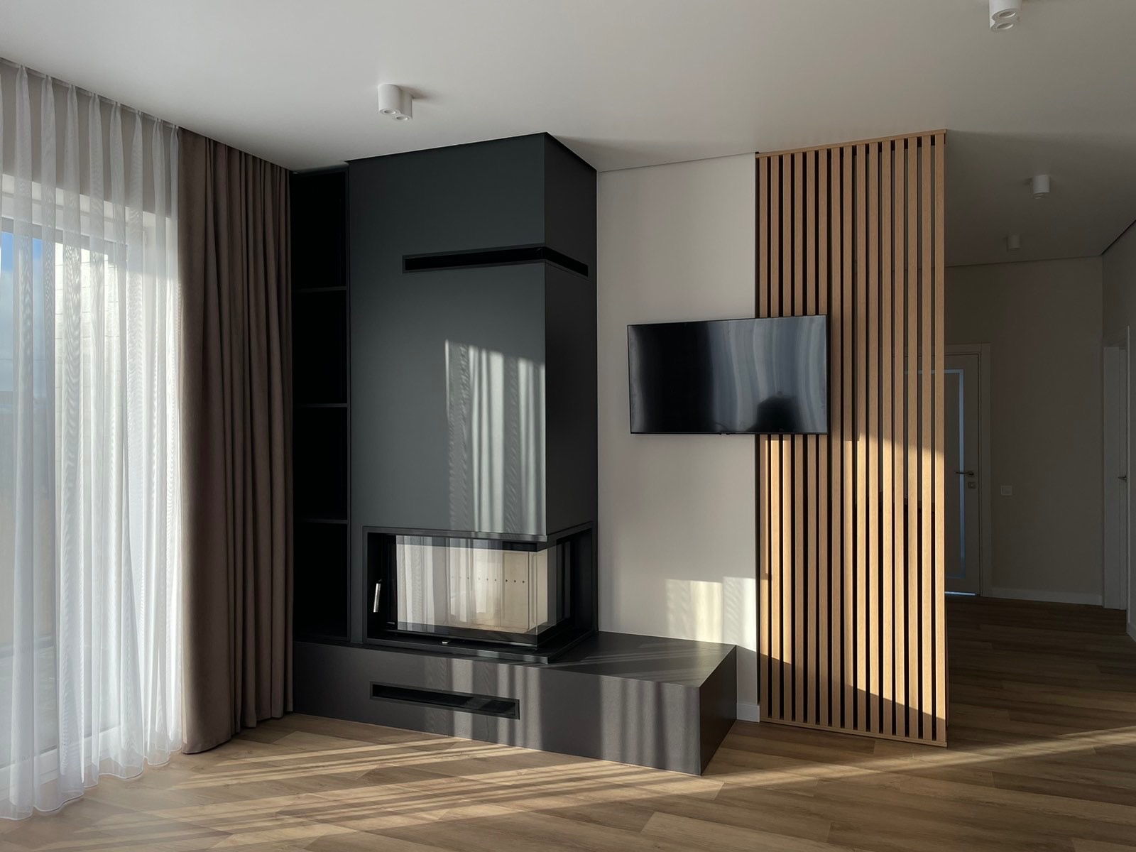 Nowoczesny salon z kominkiem o geometrycznej bryle, drewnianymi lamelami i telewizorem na ścianie. Parkiet odbija promienie słoneczne wpadające przez okno.