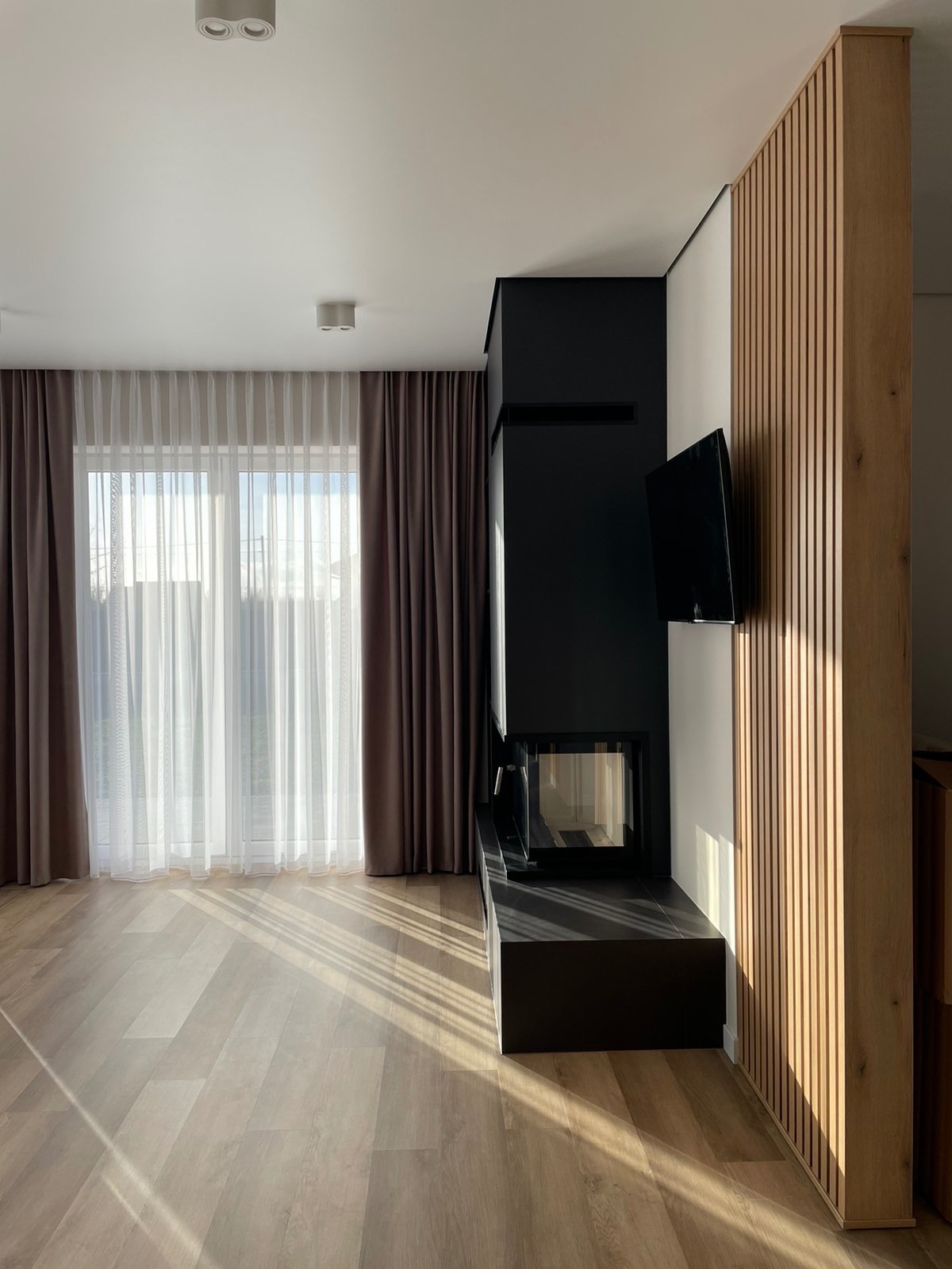Stylowe wykończenie salonu w Lublinie: drewniane lamele ścienne, czarny kominek i jasna podłoga. Nowoczesne wnętrze z zasłonami w oknie.