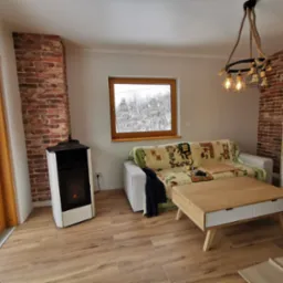 Jasne wnętrze pokoju z ceglaną ścianą, widokiem na zaśnieżone drzewa za oknem, nowoczesnym piecem na pellet, drewnianą podłogą i lampą wiszącą z grubych lin.