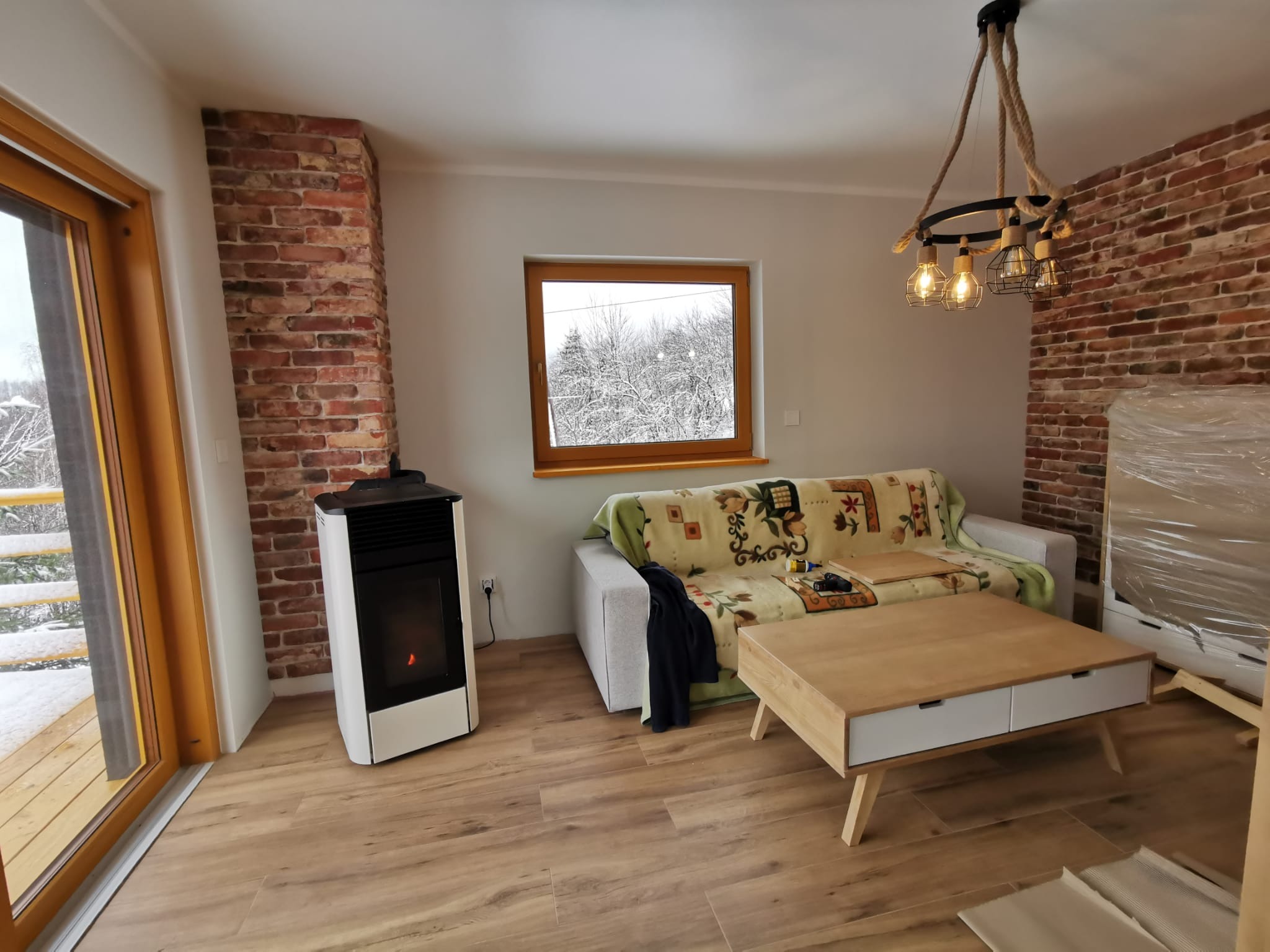 Jasne wnętrze pokoju z ceglaną ścianą, widokiem na zaśnieżone drzewa za oknem, nowoczesnym piecem na pellet, drewnianą podłogą i lampą wiszącą z grubych lin.