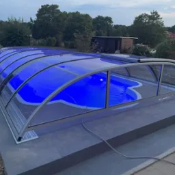 Zadaszony basen z niebieskim oświetleniem w ogrodzie, widok z góry, szara kostka brukowa wokół basenu, w tle drzewa i budynek gospodarczy, wieczorne oświetlenie.