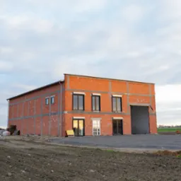 Dwukondygnacyjny budynek w trakcie budowy, z cegły i betonu komórkowego, z zamontowanymi oknami, widok na plac budowy i szklarnię w tle.