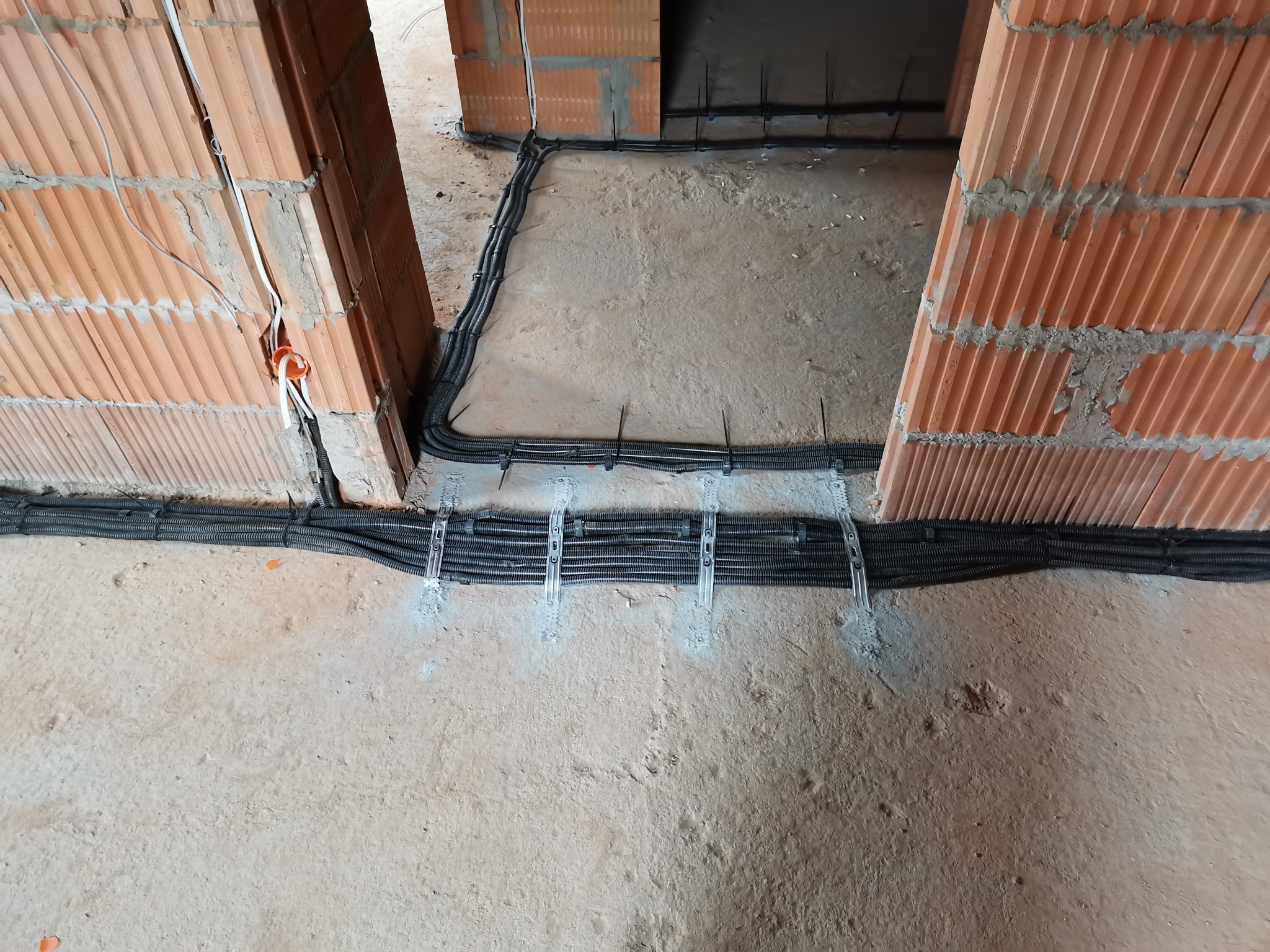 Kable elektryczne w peszlach, przymocowane do podłoża w surowym stanie budynku, widoczne ceglane ściany. Instalacja przed wylaniem posadzki.
