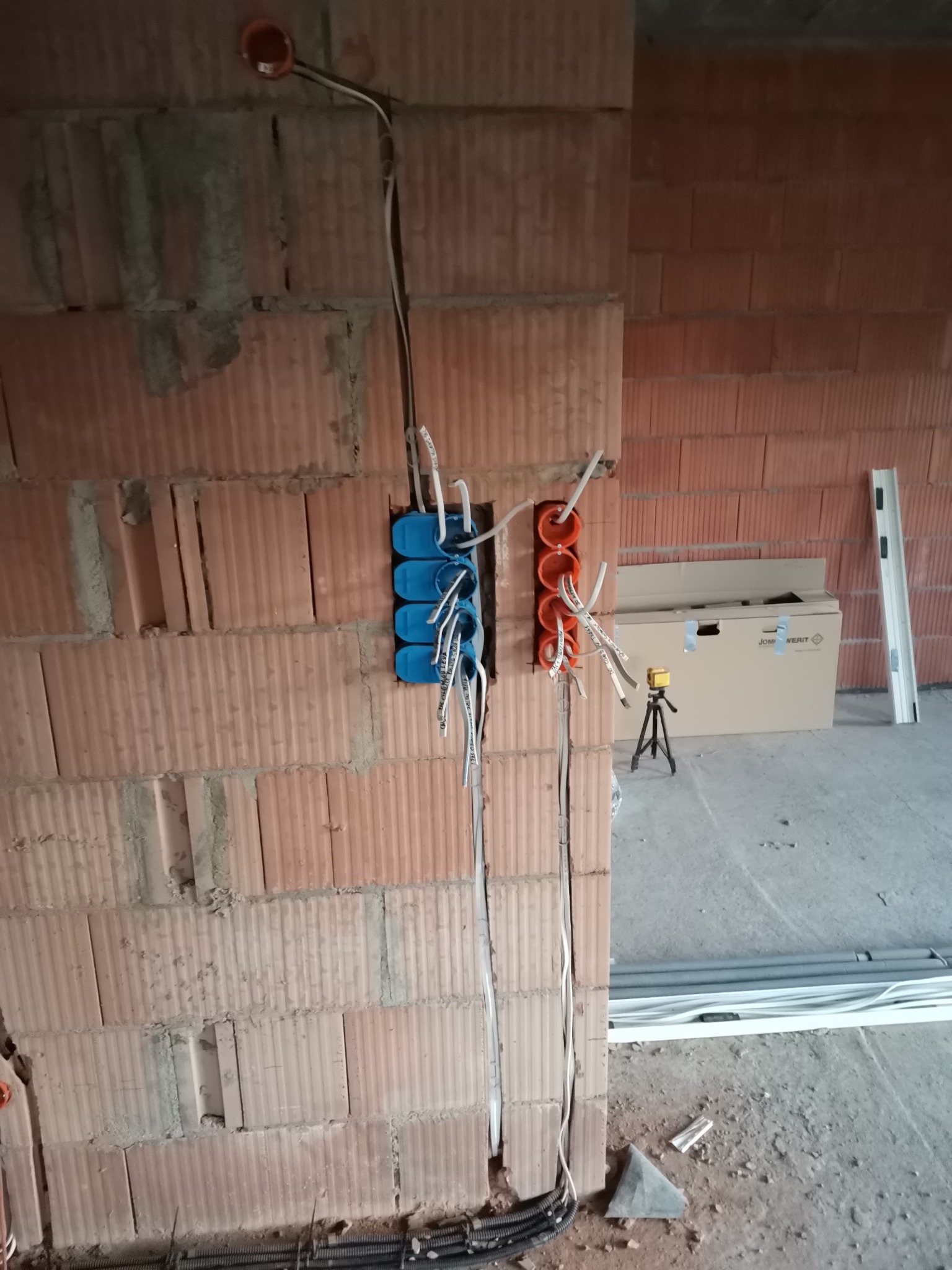 Instalacja elektryczna w trakcie montażu: kable w peszlach, puszki instalacyjne w kolorze pomarańczowym i niebieskim, ściana z czerwonej cegły dziurawki.