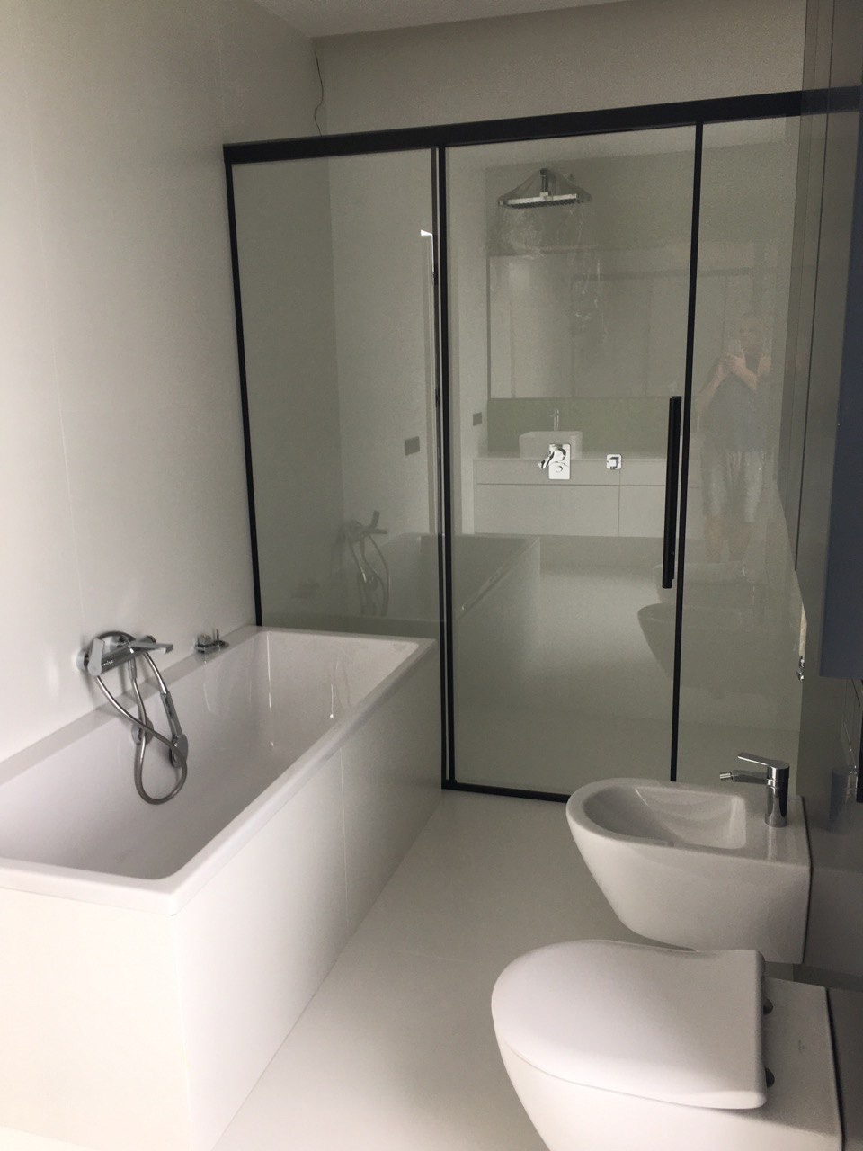 Nowoczesna, minimalistyczna łazienka z białą armaturą, kabiną prysznicową z czarną ramą i odbijającą się postacią w szkle.