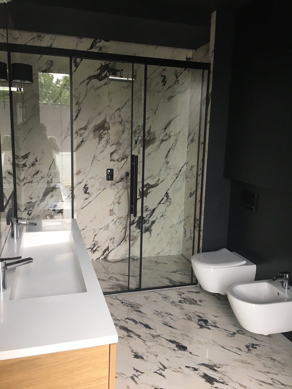 Nowoczesna łazienka w czarno-białej kolorystyce z marmurowymi płytkami, kabiną prysznicową typu walk-in z czarnymi profilami, wiszącą toaletą i bidetem oraz umywalką nablatową na drewnianej szafce.