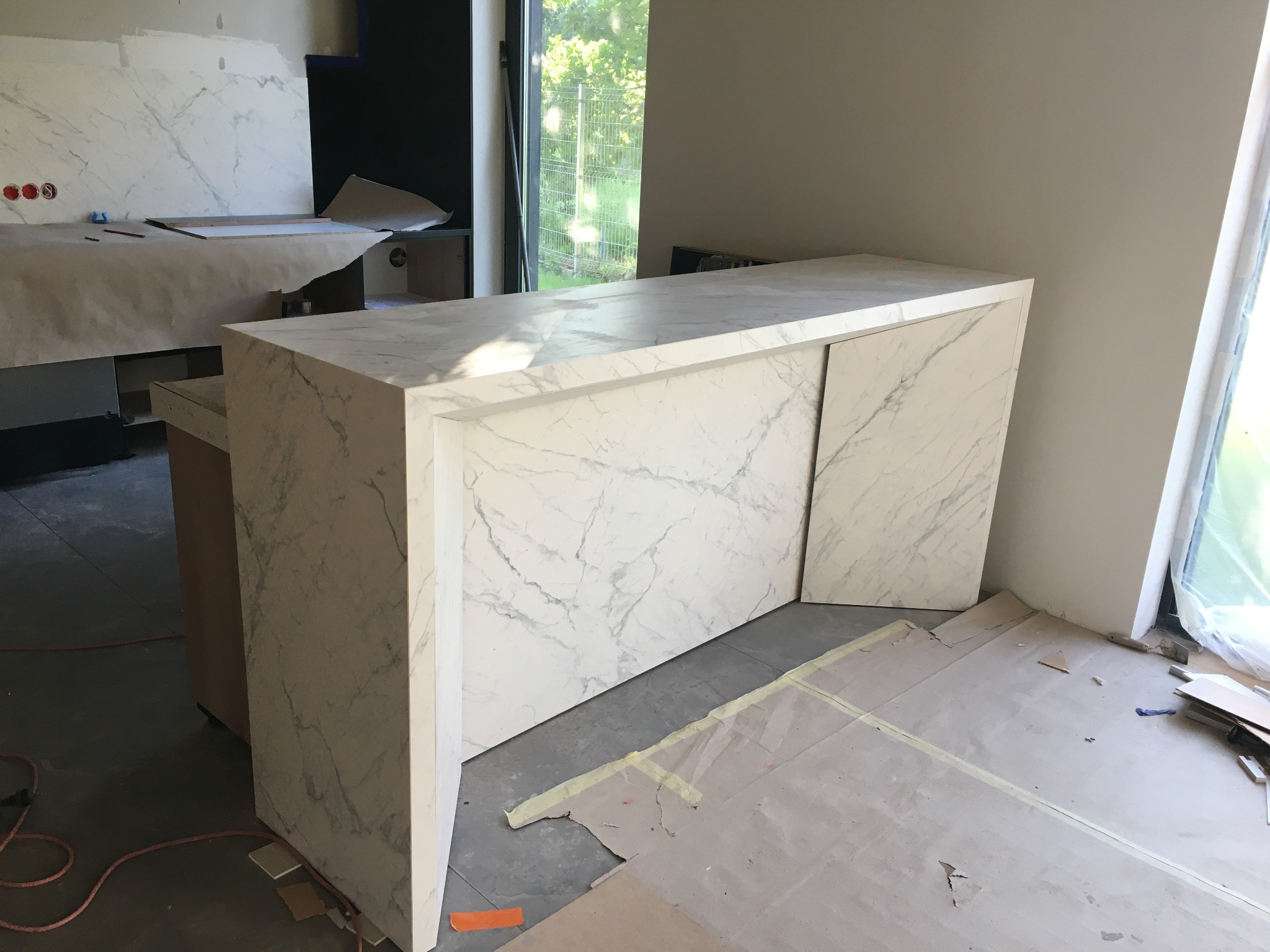 Biała wyspa kuchenna z marmurowym wzorem, zamontowana w surowym wnętrzu, z zabezpieczoną podłogą i panelami ściennymi w tle.