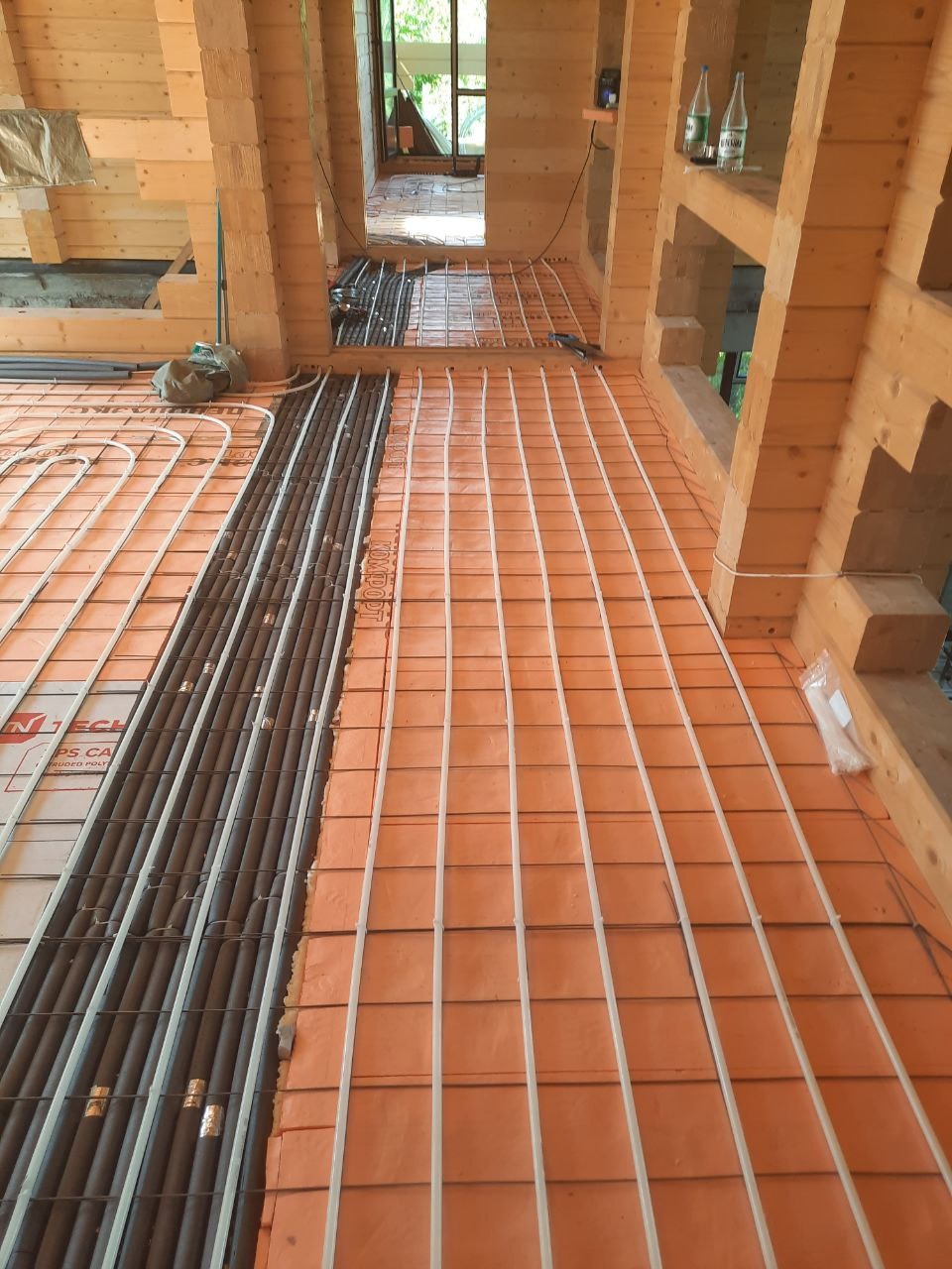 Montaż ogrzewania podłogowego: rury grzewcze ułożone na pomarańczowej izolacji termicznej, widoczne drewniane elementy konstrukcyjne domu w surowym stanie.