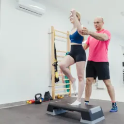 Kobieta w sportowym stroju ćwiczy z trenerem na stepie w jasnym gabinecie, lustro odbija jej sylwetkę, widoczne drabinki gimnastyczne i sprzęt sportowy w tle.