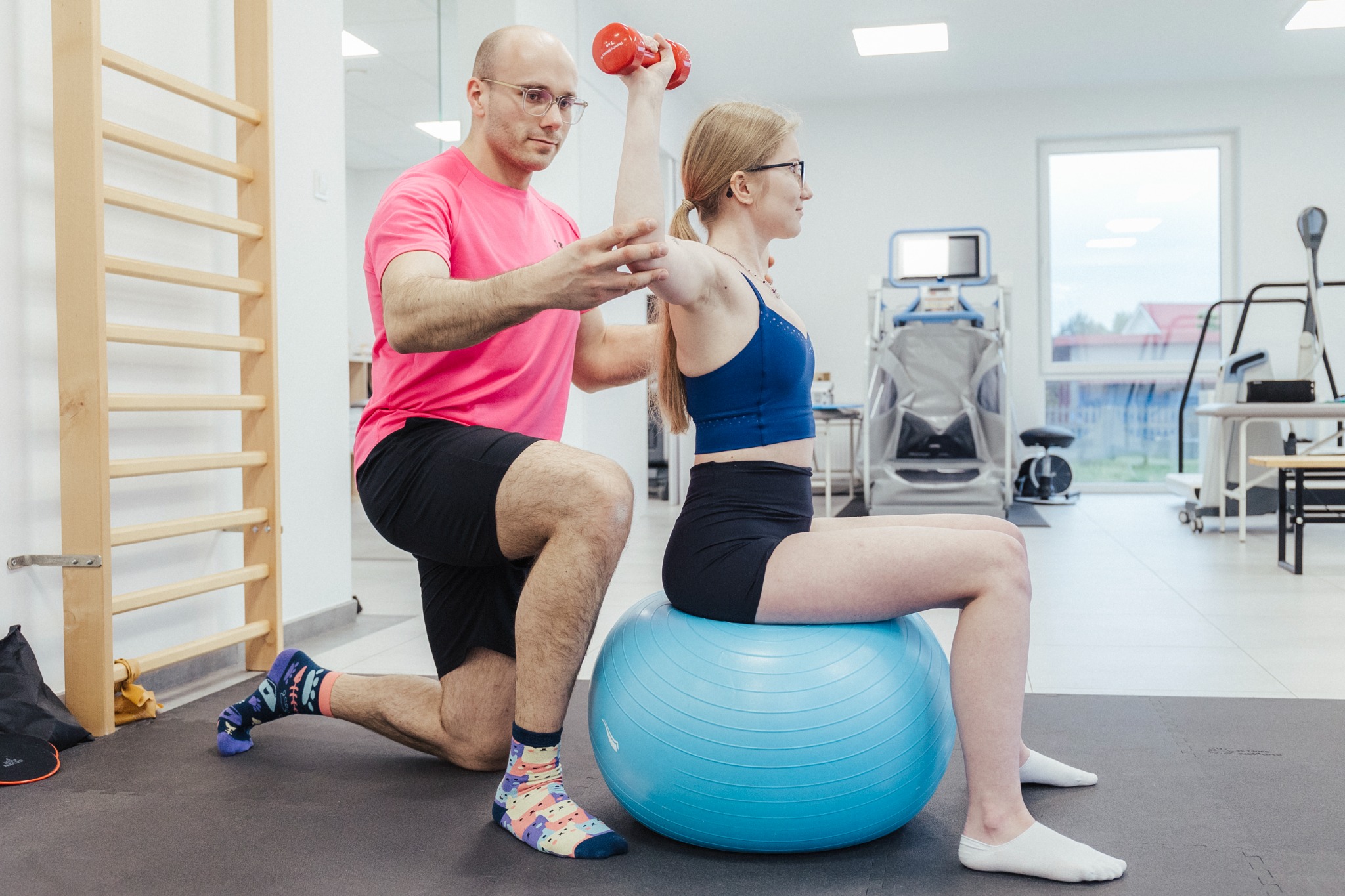 Kobieta w okularach siedzi na niebieskiej piłce gimnastycznej, unosząc czerwoną hantlę, podczas gdy mężczyzna w różowej koszulce i wzorzystych skarpetkach stabilizuje jej barki w jasnym gabinecie...