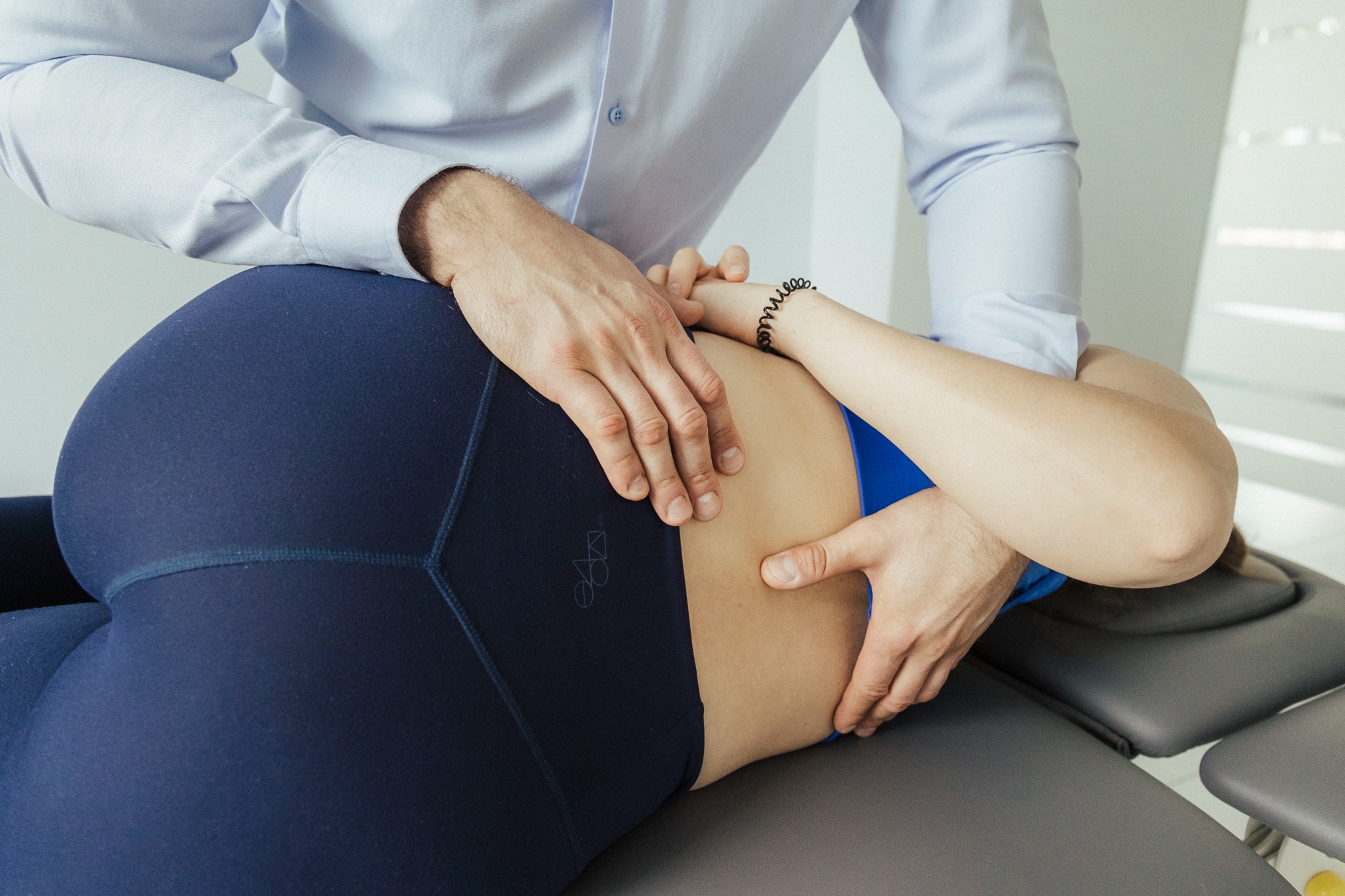 Pacjentka w granatowych legginsach leży na szarym łóżku rehabilitacyjnym, terapeuta w jasnoniebieskiej koszuli uciska jej dolną część pleców obiema dłońmi.
