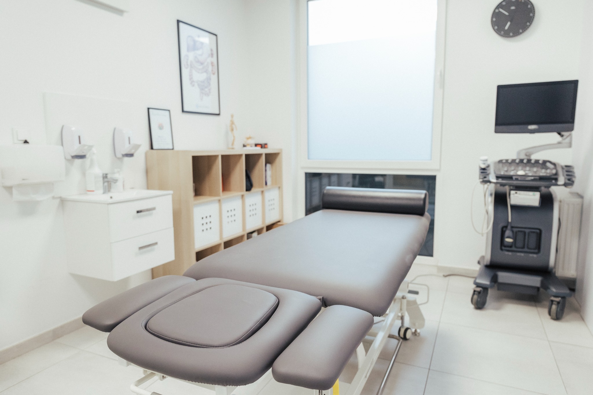 Jasne wnętrze gabinetu z szarym, regulowanym łóżkiem do zabiegów, szafką z umywalką, regałem z koszykami i aparaturą diagnostyczną z monitorem.