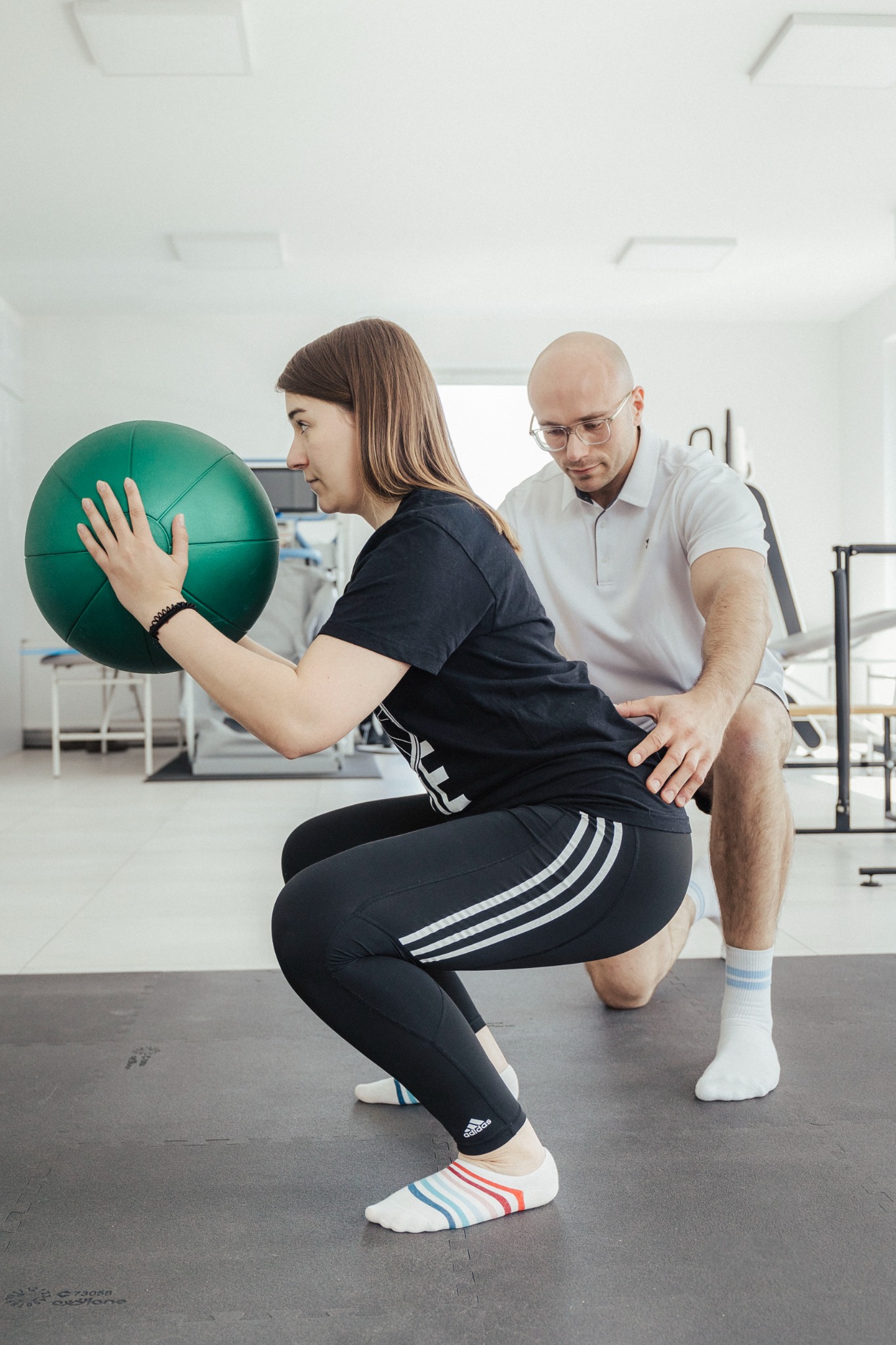 Kobieta w czarnych legginsach z trzema paskami ćwiczy przysiad z zieloną piłką, a mężczyzna w białej koszulce i okularach koryguje jej postawę, stojąc na kolanach.