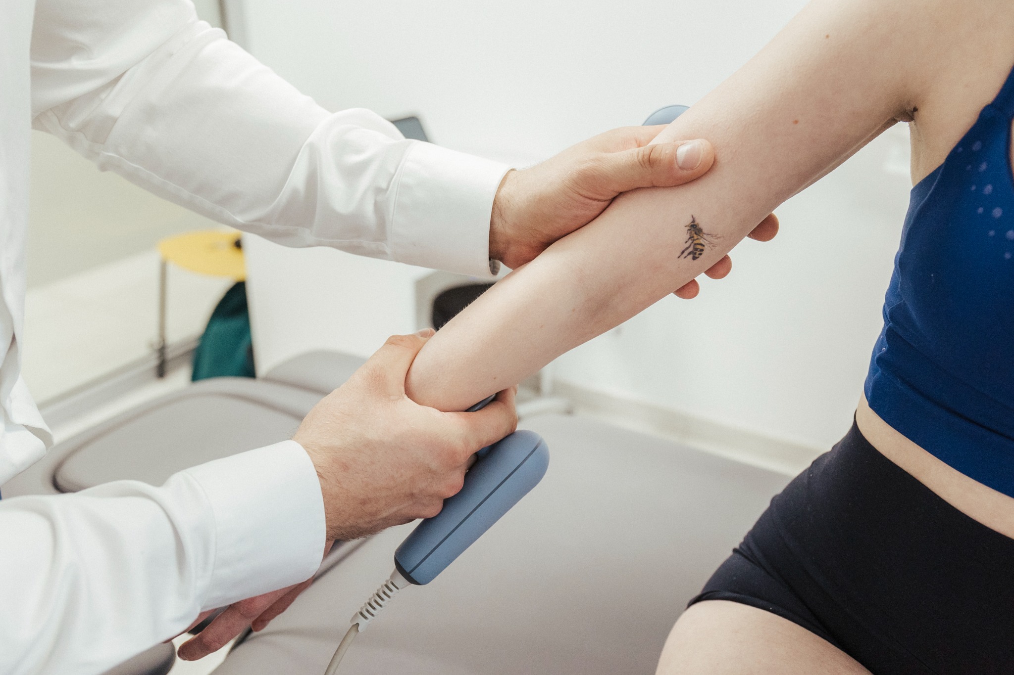 Pacjentka w sportowym stroju podczas zabiegu z użyciem urządzenia do fizykoterapii na ramieniu z tatuażem pszczoły, wykonywanego przez osobę w białym fartuchu na tle białego kozetki.