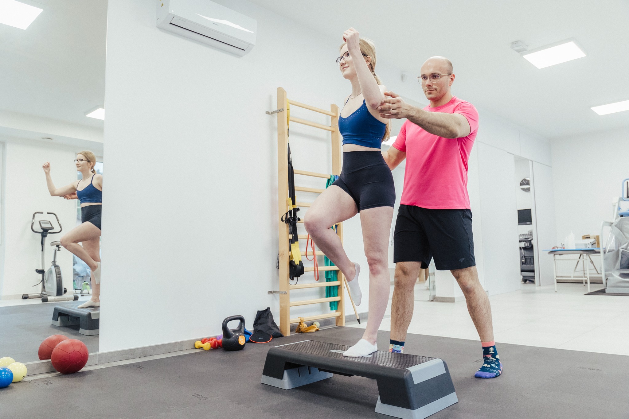 Kobieta w sportowym stroju ćwiczy z trenerem na stepie w jasnym gabinecie, lustro odbija jej sylwetkę, widoczne drabinki gimnastyczne i sprzęt sportowy w tle.