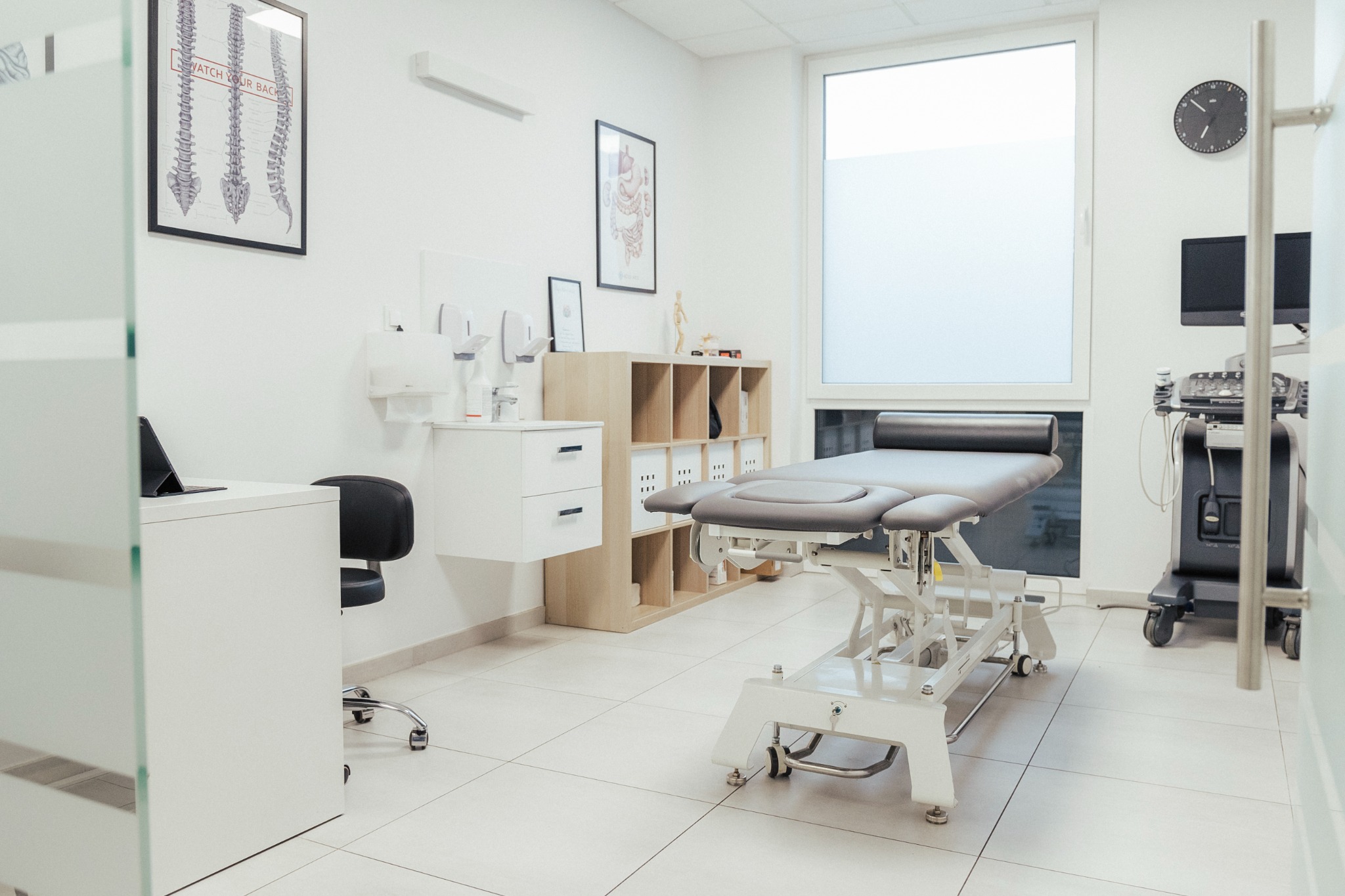 Jasne wnętrze gabinetu z profesjonalnym stołem do terapii, biurkiem z tabletem, szafką z przyborami i plakatem z kręgosłupem na ścianie.