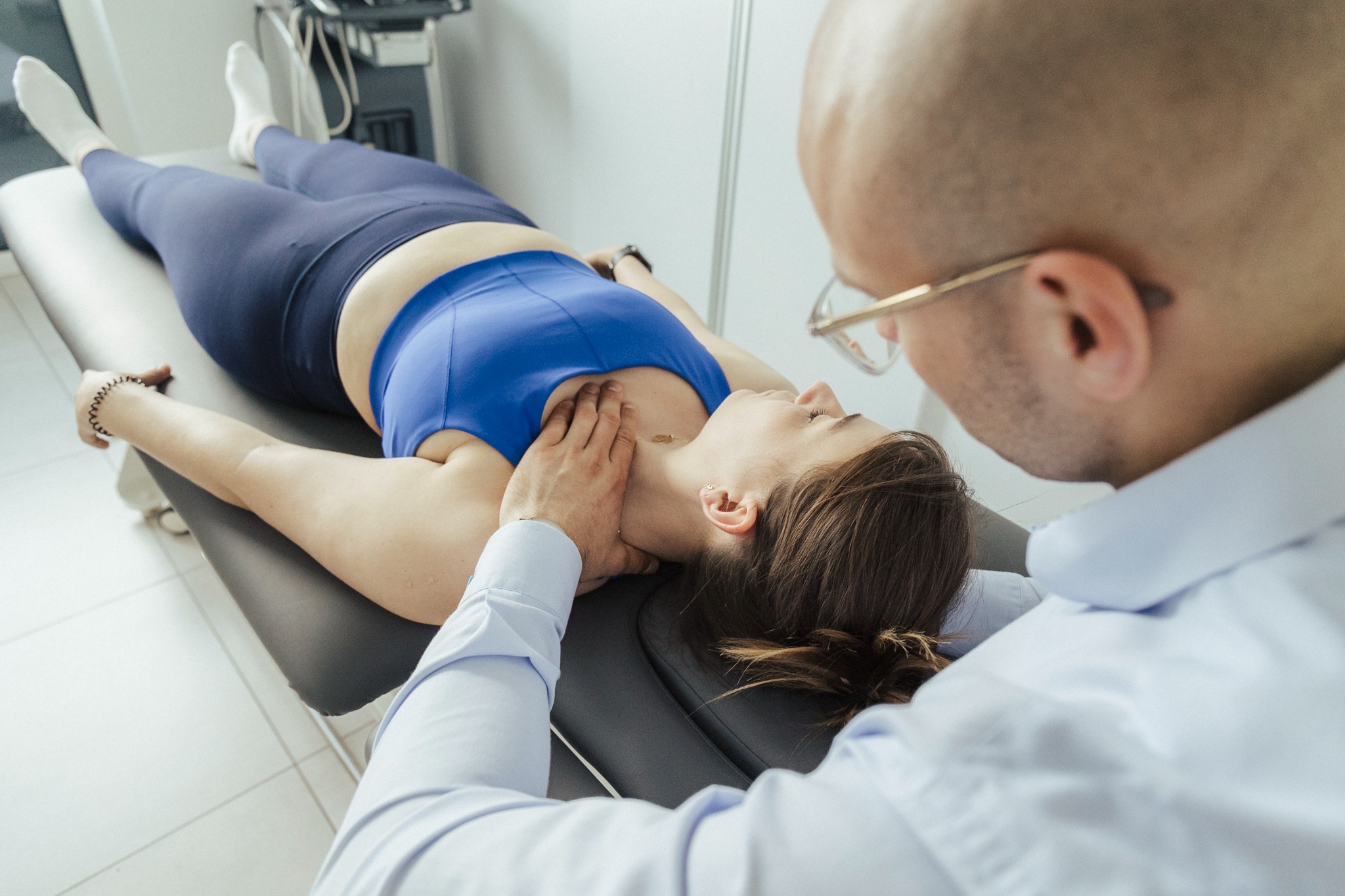 Kobieta leżąca na kozetce podczas badania palpacyjnego szyi przez mężczyznę w okularach i białej koszuli, z urządzeniem diagnostycznym w tle.