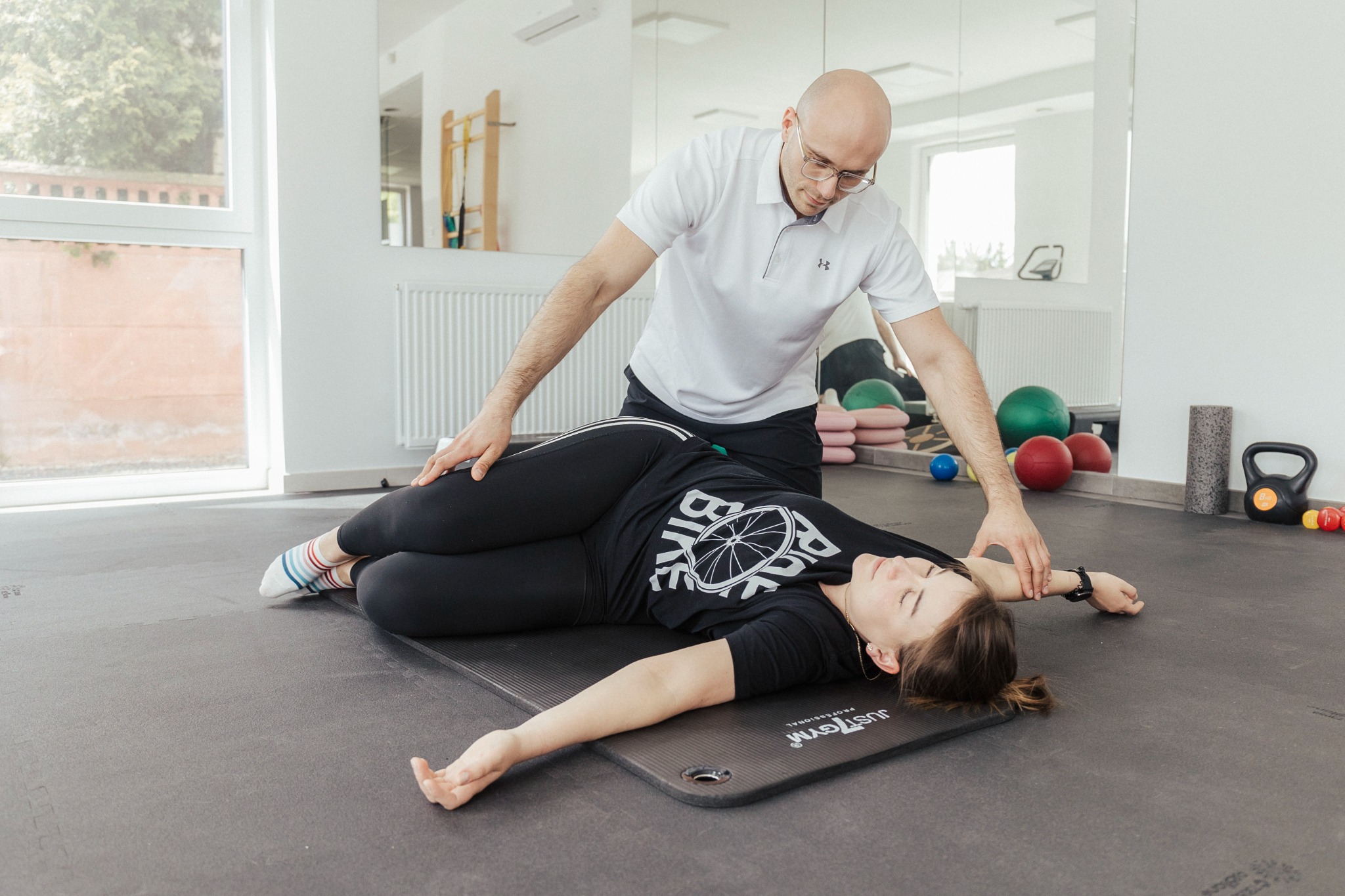 Kobieta leżąca na macie do ćwiczeń podczas sesji terapeutycznej, terapeuta w białej koszulce delikatnie koryguje ułożenie jej ramienia, w tle lustro i sprzęt do ćwiczeń.