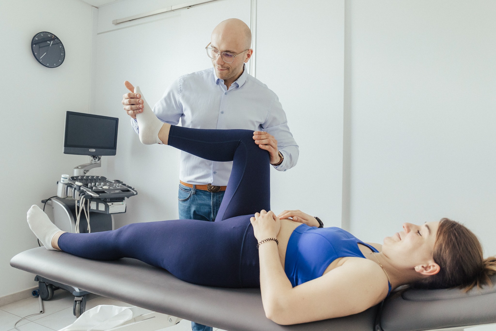 Pacjentka leży na kozetce, a specjalista w okularach delikatnie podnosi jej nogę w białej skarpetce, wykonując ćwiczenie rozciągające; w tle widoczny aparat USG i zegar na ścianie.