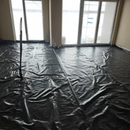 Przygotowanie do wylewki: czarna folia ochronna rozłożona na podłodze w pomieszczeniu z oknami, widoczny wężyk instalacji ogrzewania podłogowego.