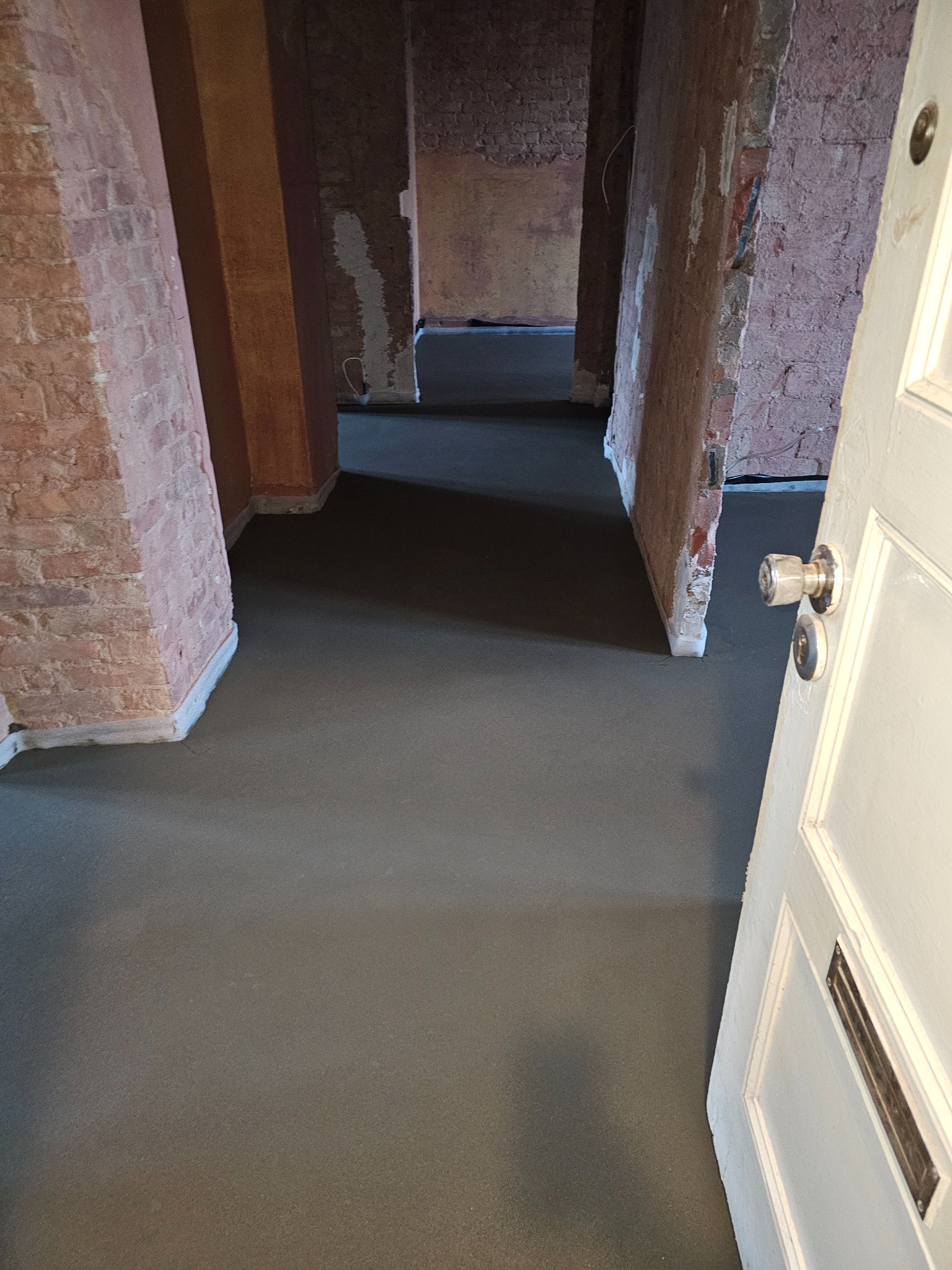 Świeżo wylana, gładka posadzka jastrychowa w pomieszczeniu z odsłoniętą cegłą i otwartymi drzwiami, widok z perspektywy wejścia.