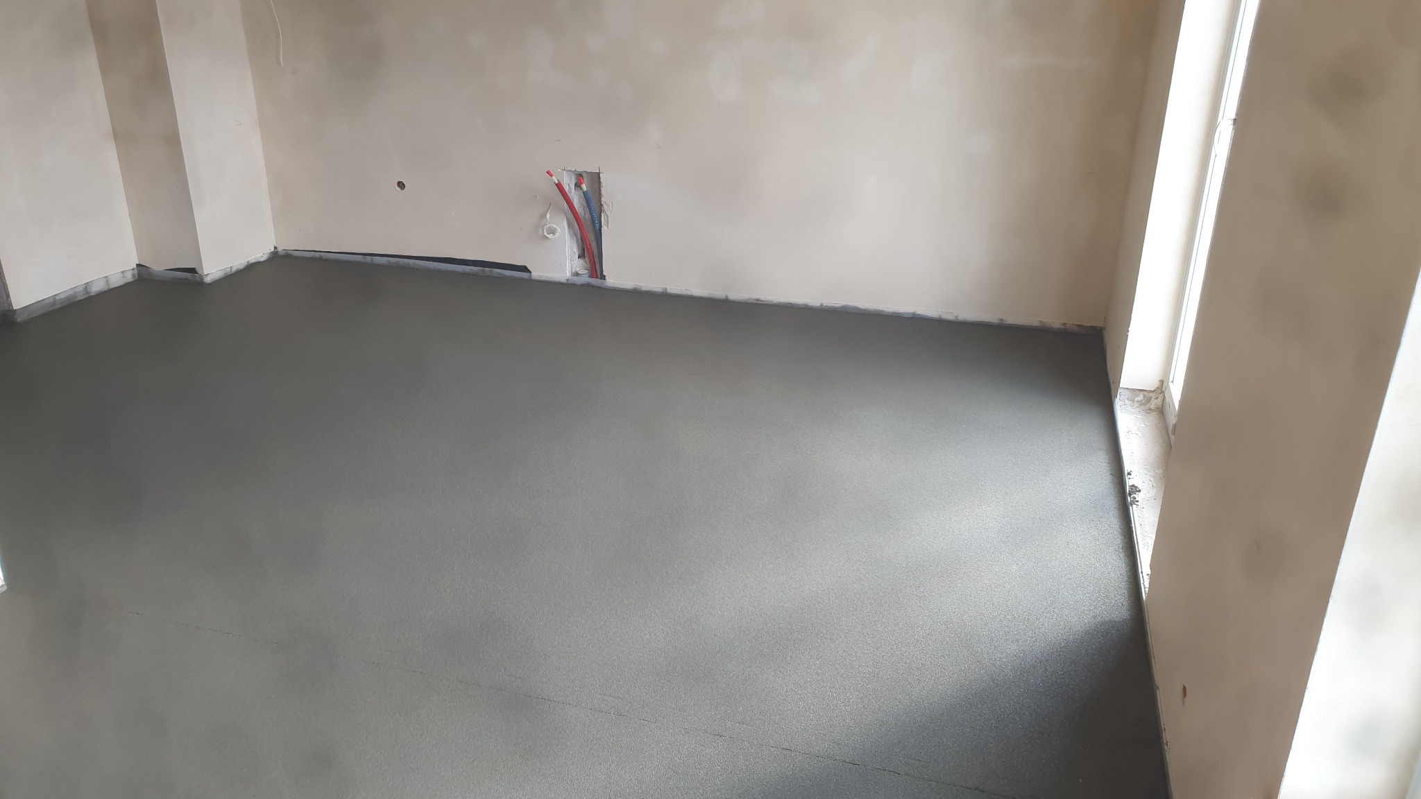 Świeżo wylana, gładka posadzka w jasnym pomieszczeniu z widocznymi rurami instalacji w ścianie.