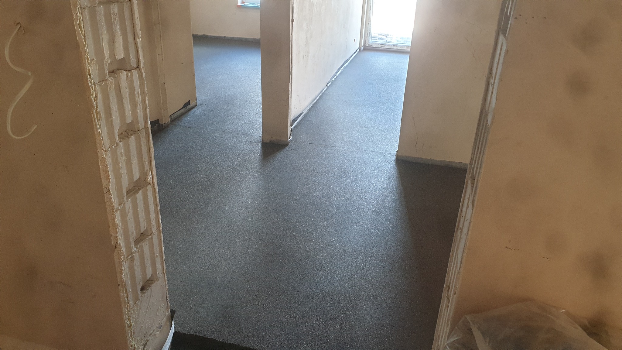 Świeżo wylana, gładka, szara posadzka w pomieszczeniu z surowymi ścianami i filarami, widok z perspektywy wejścia.