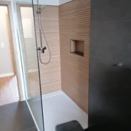 Nowoczesna kabina prysznicowa z deszczownicą, wykończona drewnopodobnymi panelami i ciemnymi płytkami, z wnęką w ścianie i szarą wycieraczką na podłodze z ciemnych płytek.