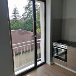 Wnętrze kuchni z widokiem na balkon i zieleń za oknem, nowoczesna zabudowa kuchenna z płytą grzewczą i piekarnikiem.