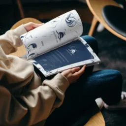 Osoba siedząca w fotelu przegląda menu hotelu Łeba & Spa, z widocznym logo i informacjami o godzinach otwarcia restauracji. Menu utrzymane w kolorystyce bieli i granatu, z elementami graficznymi...