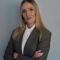 Portret kobiety w szarej marynarce i białej bluzce, z założonymi rękami na tle szarej ściany. Subtelny makijaż i blond włosy. Uśmiech.