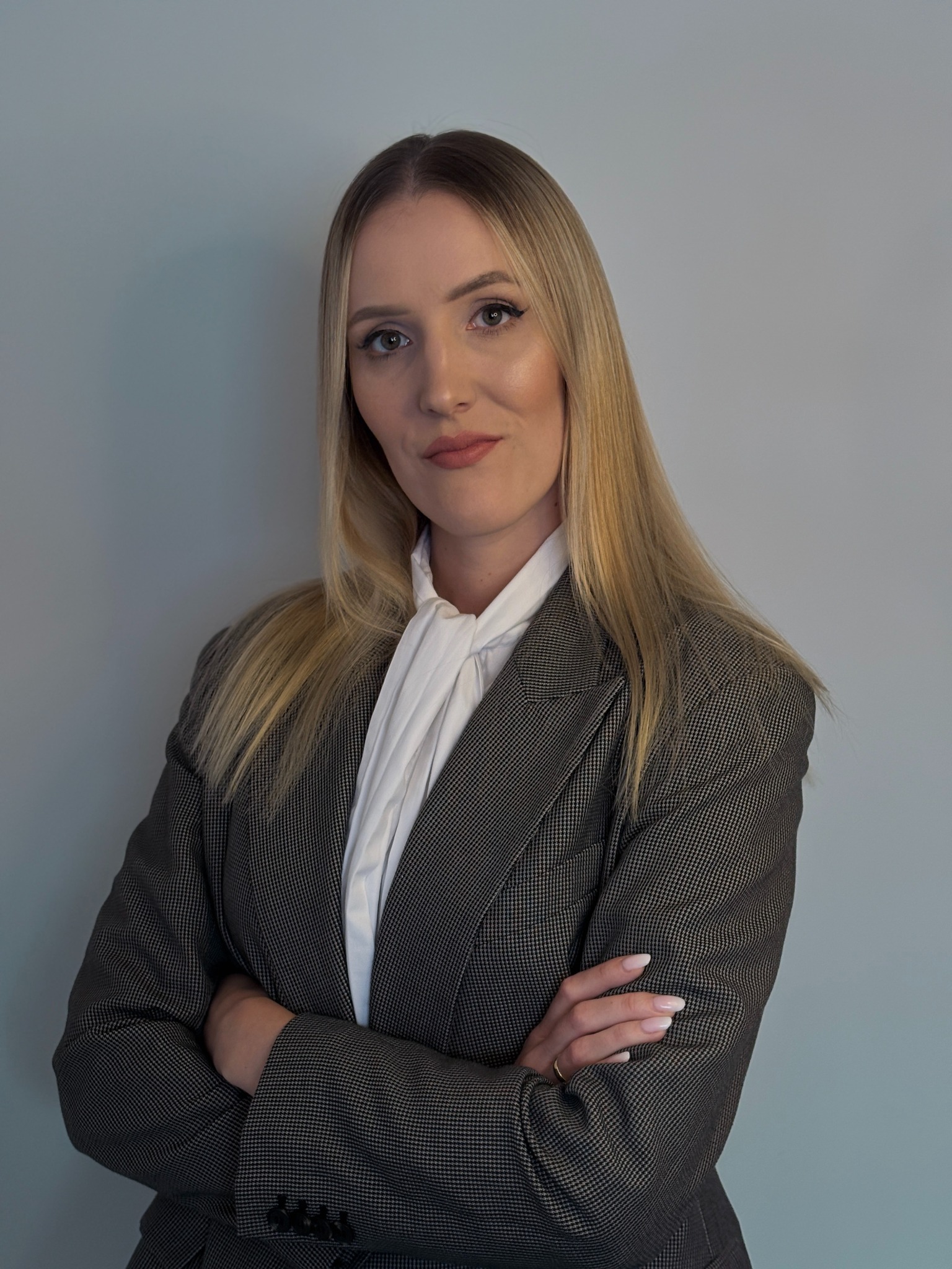 Portret kobiety w szarej marynarce i białej bluzce, z założonymi rękami na tle szarej ściany. Subtelny makijaż i blond włosy. Uśmiech.