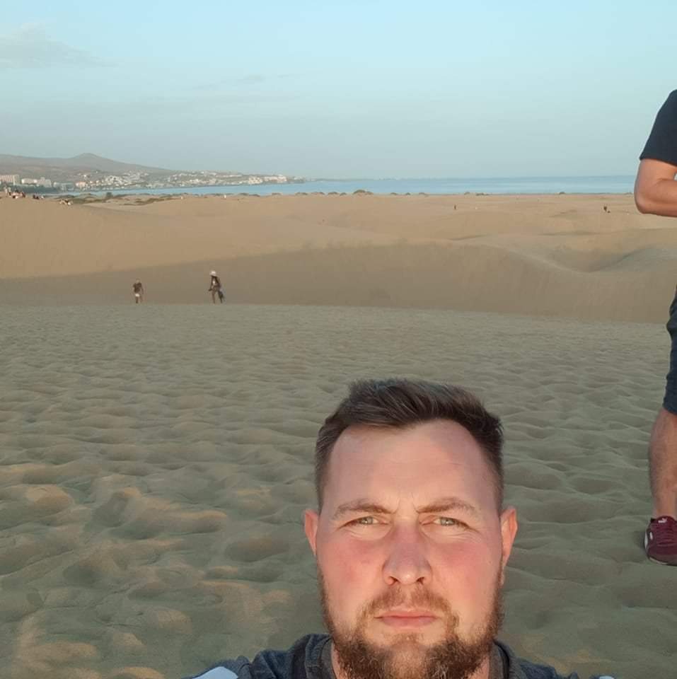 Selfie mężczyzny z brodą na tle piaszczystych wydm, z widocznym wybrzeżem i budynkami w oddali, oraz fragmentem sylwetki innej osoby po prawej stronie kadru.