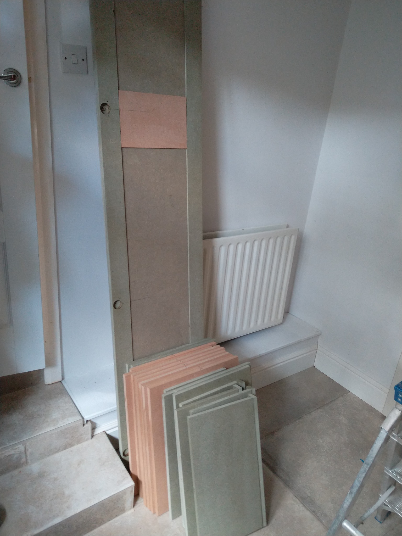 Przygotowane panele podłogowe w odcieniach beżu i szarości, ułożone obok drzwi z widocznymi otworami na klamkę i zamki, w tle biały grzejnik i fragment drabiny.