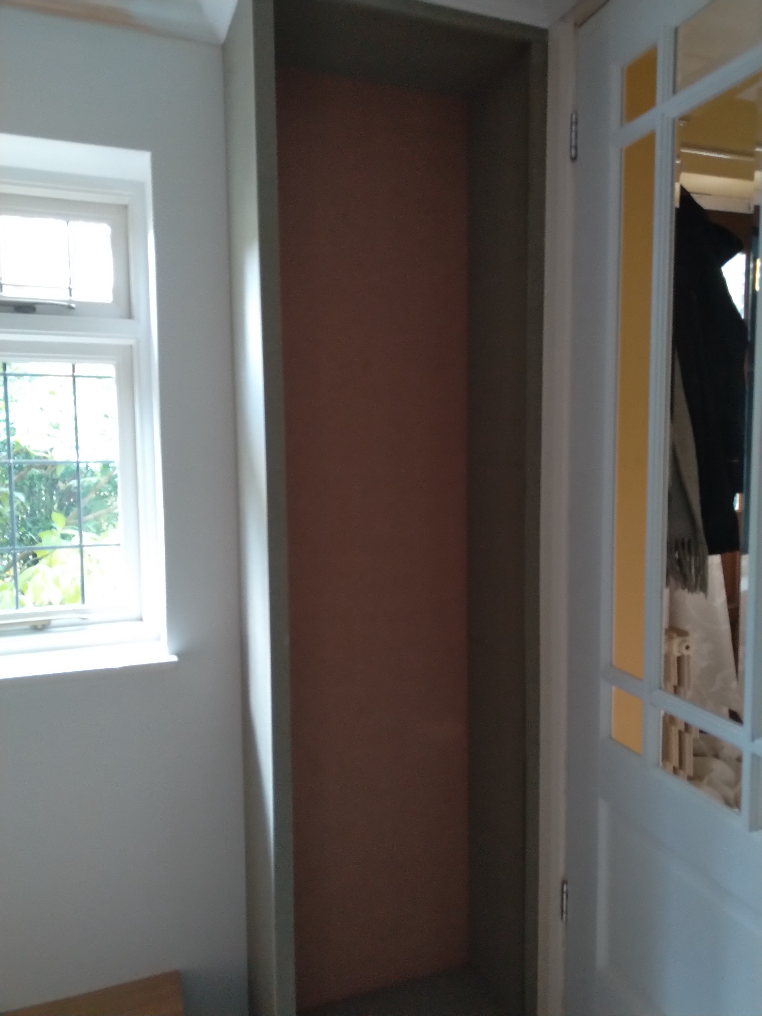 Wnęka w ścianie przed malowaniem, widoczne ślady po szpachlowaniu i przygotowaniu do nałożenia farby, obok okno z widokiem na zieleń i fragment drzwi z szybkami.