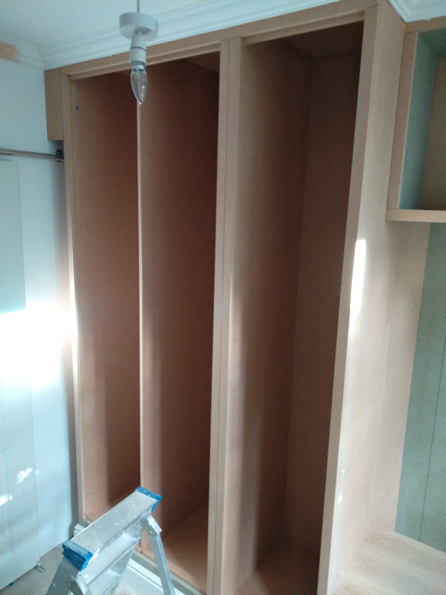 Montaż szafy wnękowej z surowej płyty MDF, widoczna drabina aluminiowa i żarówka wisząca na kablu.