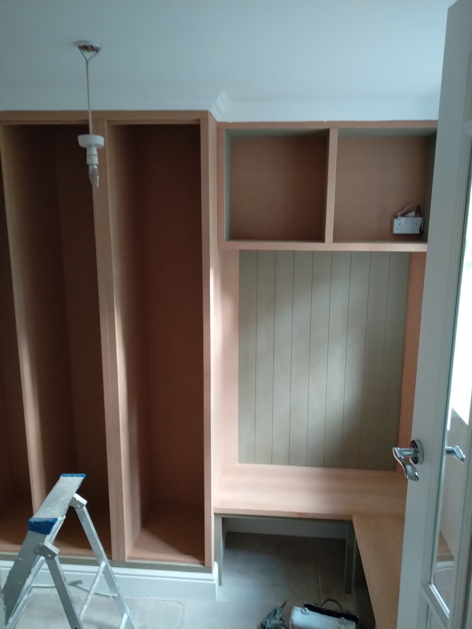 Wnętrze w trakcie tworzenia zabudowy meblowej z drewnianymi półkami i panelami ściennymi, widoczna drabina i narzędzia.