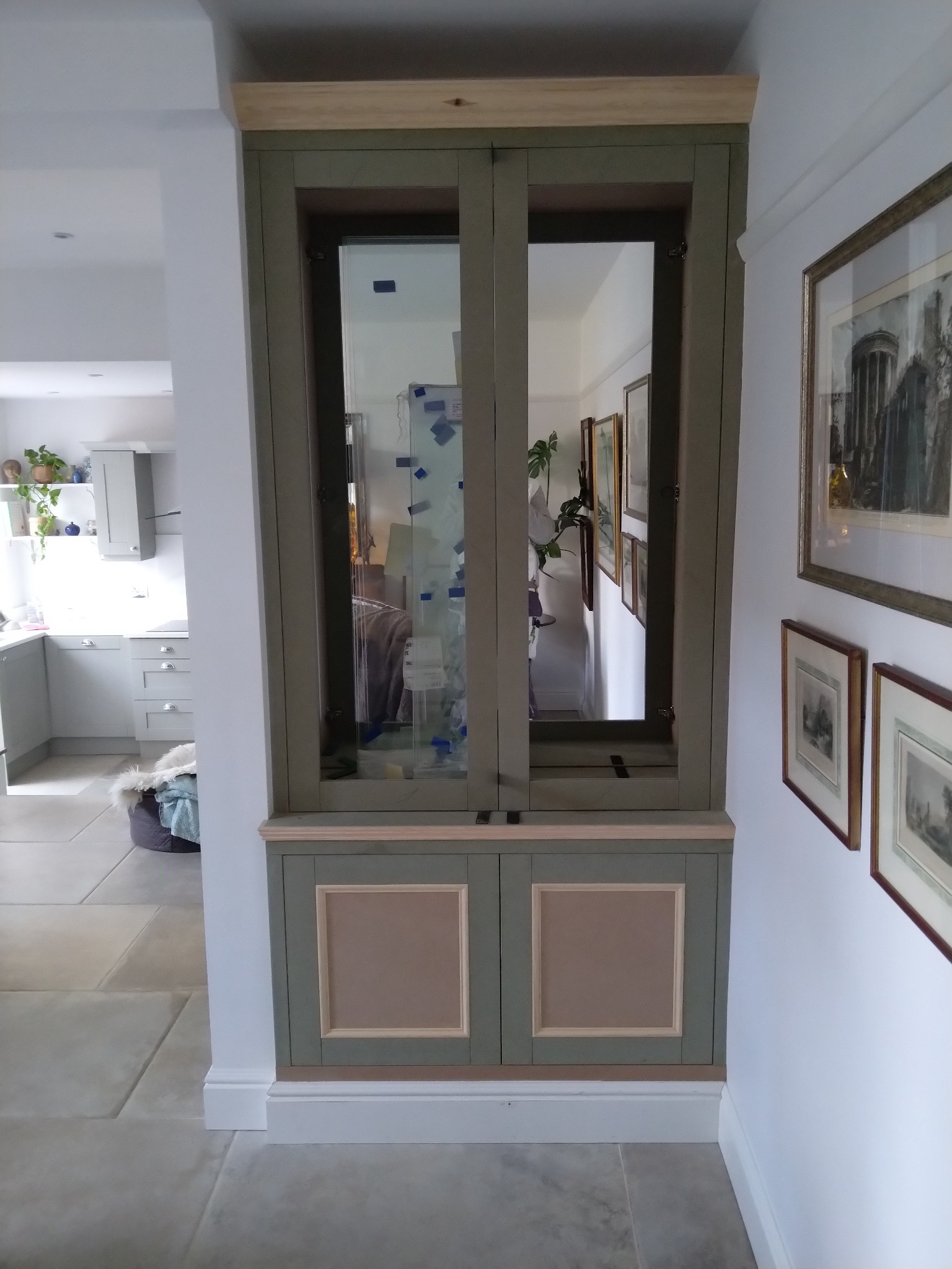 Wbudowana szafka z przeszklonymi drzwiami w odcieniach zieleni, z widocznymi elementami wykończeniowymi w surowym drewnie, wkomponowana w ścianę między kuchnią a korytarzem.