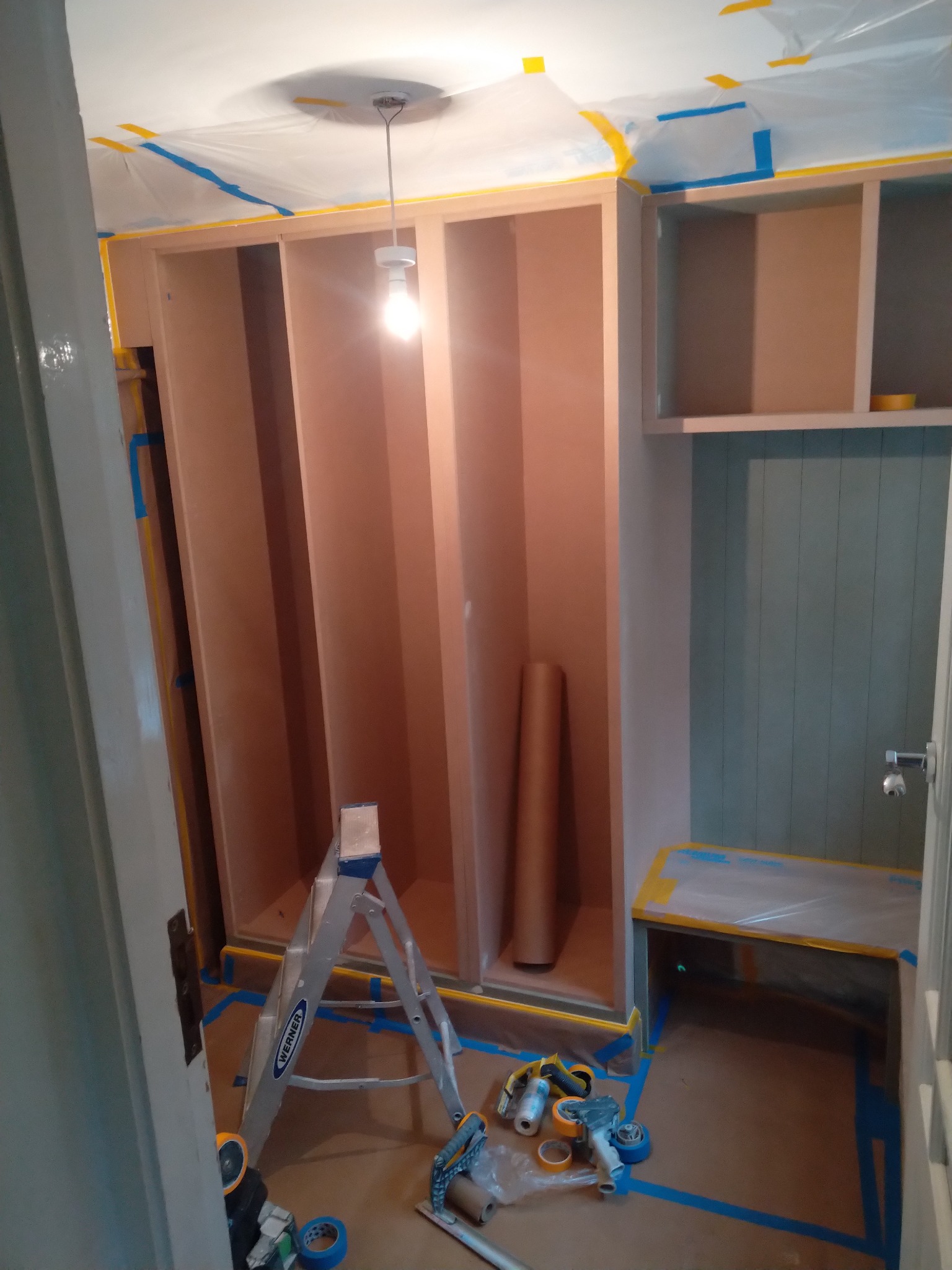 Wnętrze pokoju w trakcie przygotowań do prac wykończeniowych: zabezpieczone folią i taśmą malarską ściany, sufit i meble, rozstawiona drabina, widoczny wałek malarski i taśmy.