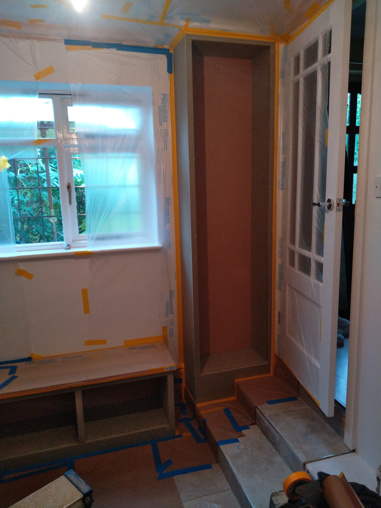 Przygotowane do malowania wnętrze z zabezpieczonymi folią i taśmą meblami, oknem i drzwiami, widoczne schody wyłożone płytkami.