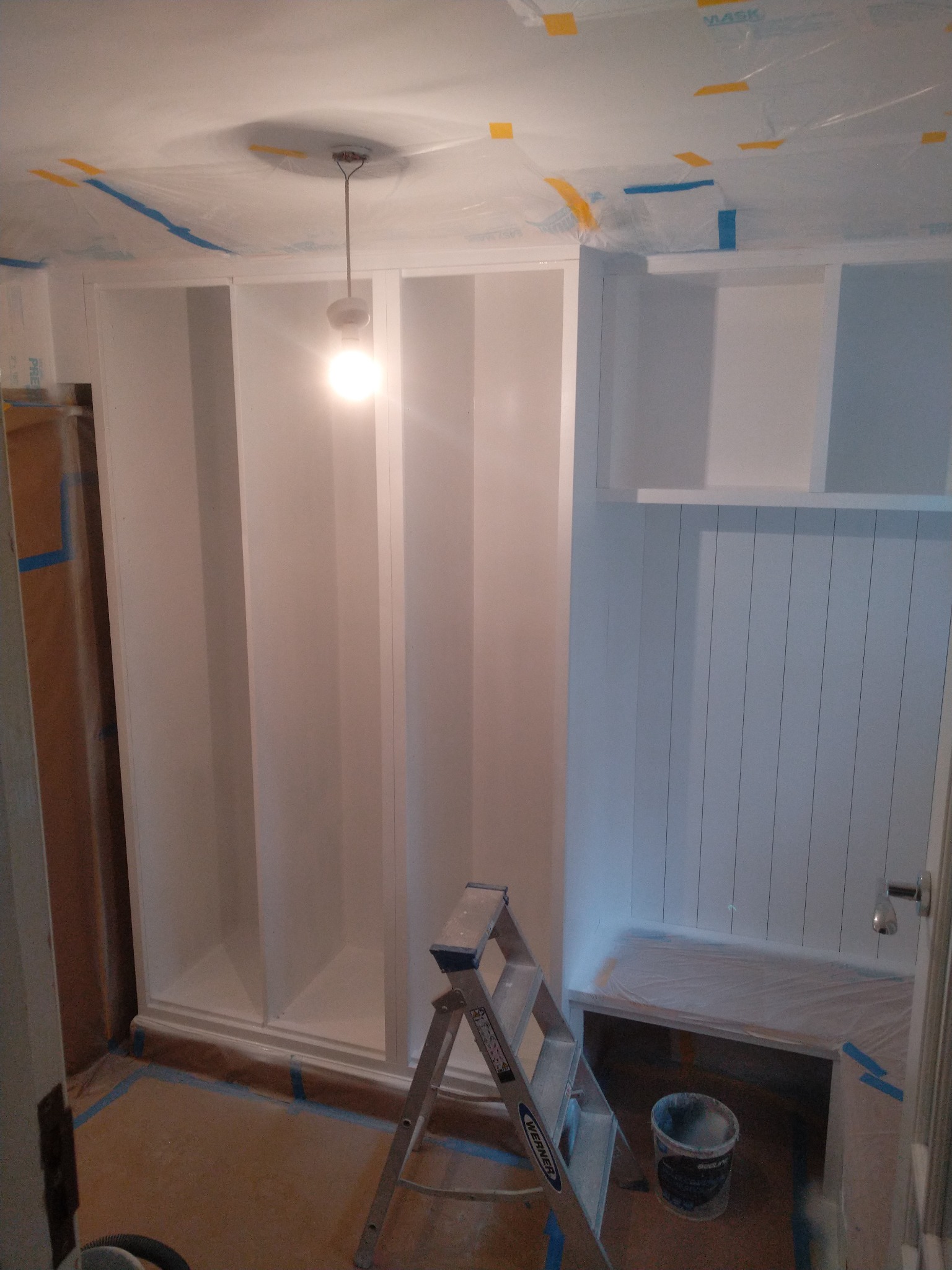 Białe, nowo pomalowane regały wnękowe z otwartymi półkami, zabezpieczone folią malarską podłogi i sufit, drabina malarska i wiadro z farbą w pomieszczeniu w trakcie prac wykończeniowych.