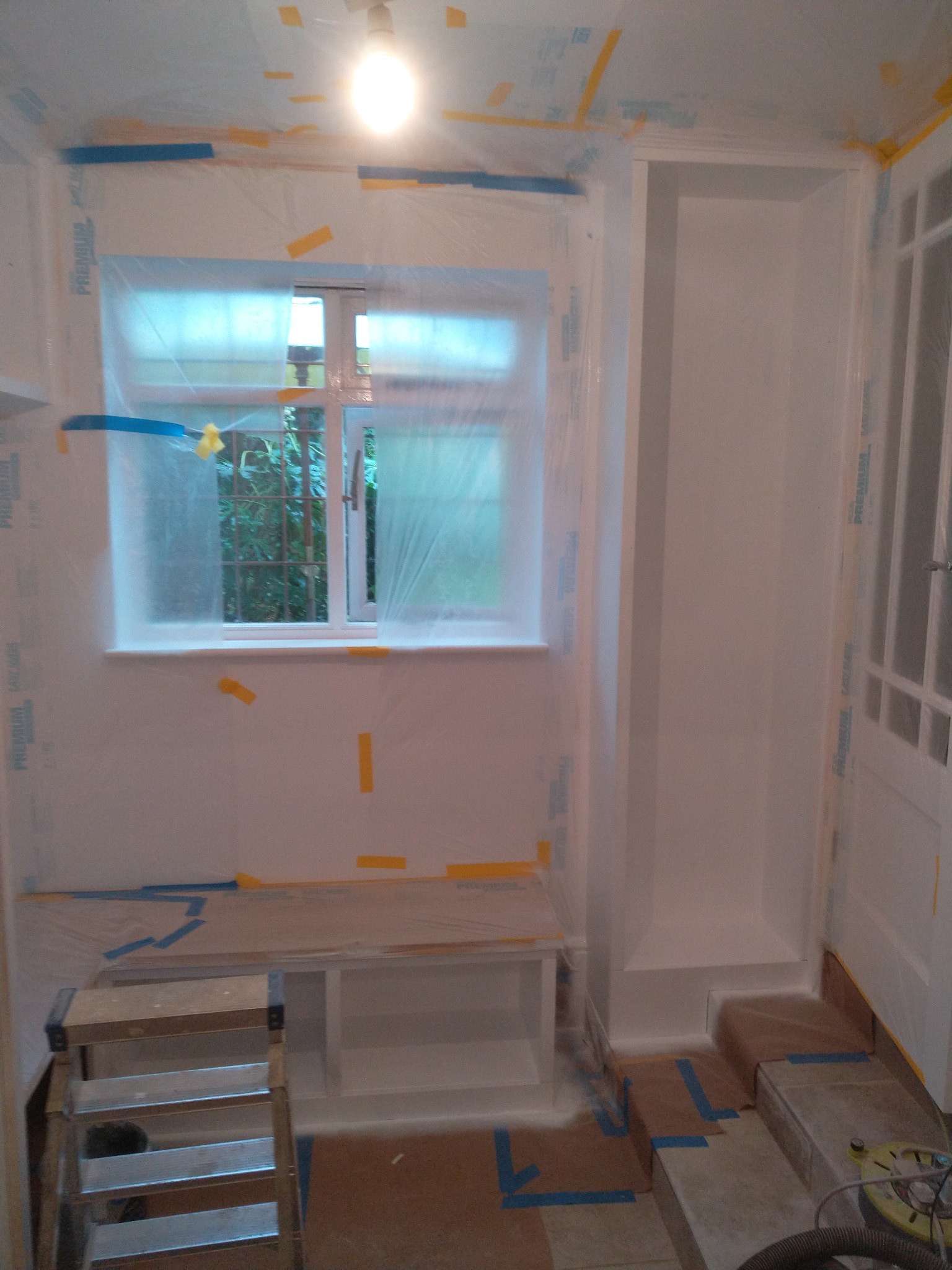 Pokój zabezpieczony folią i taśmą malarską przed malowaniem, widoczna drabina, okno z widokiem na zieleń, wnęka na szafę i częściowo zakryte podłogi.