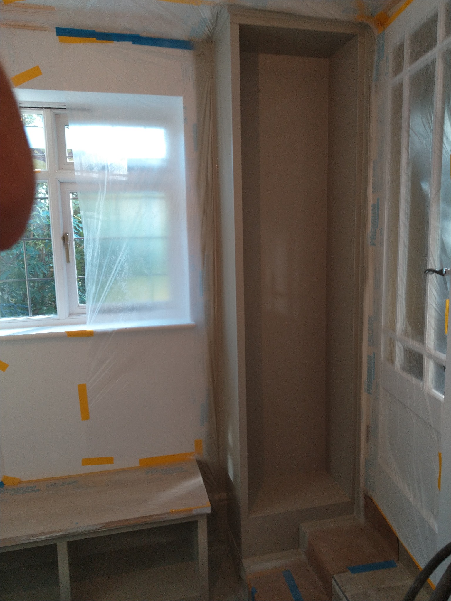 Przygotowanie do malowania pokoju: okno i drzwi zabezpieczone folią malarską, ściany oklejone taśmą, wnęka w ścianie przygotowana do malowania.