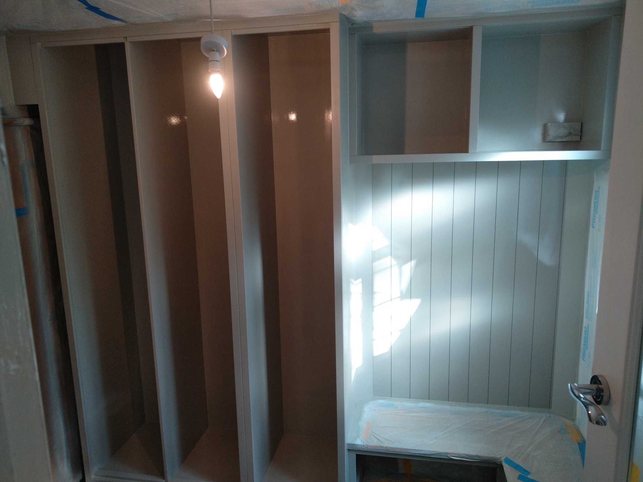 Wnętrze szafy wnękowej w trakcie montażu, widoczne pionowe przegrody, półki i siedzisko zabezpieczone folią ochronną, oświetlone wiszącą żarówką