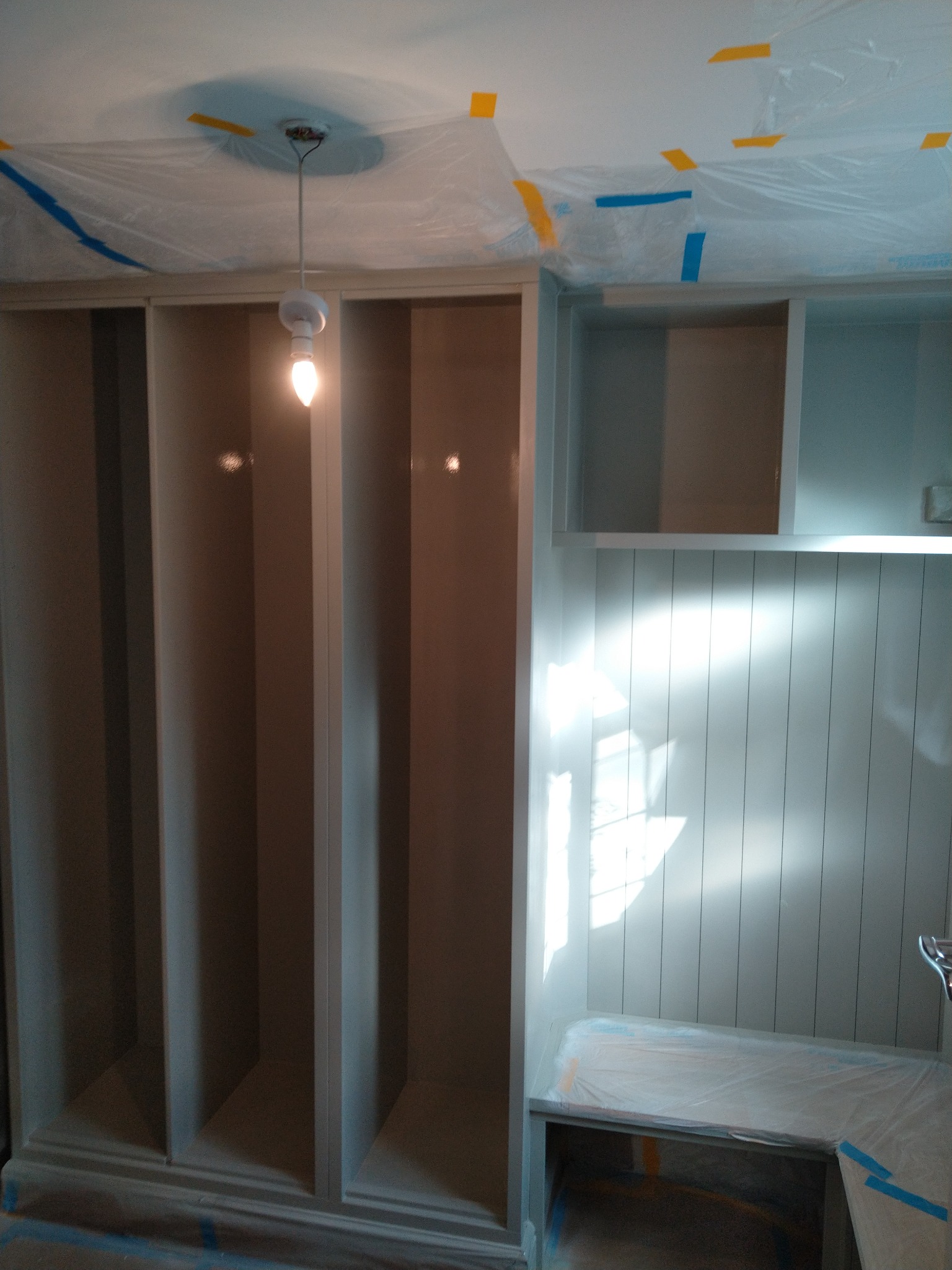 Wnęka z półkami i siedziskiem w trakcie odświeżania, zabezpieczona folią malarską, widoczna surowa żarówka.